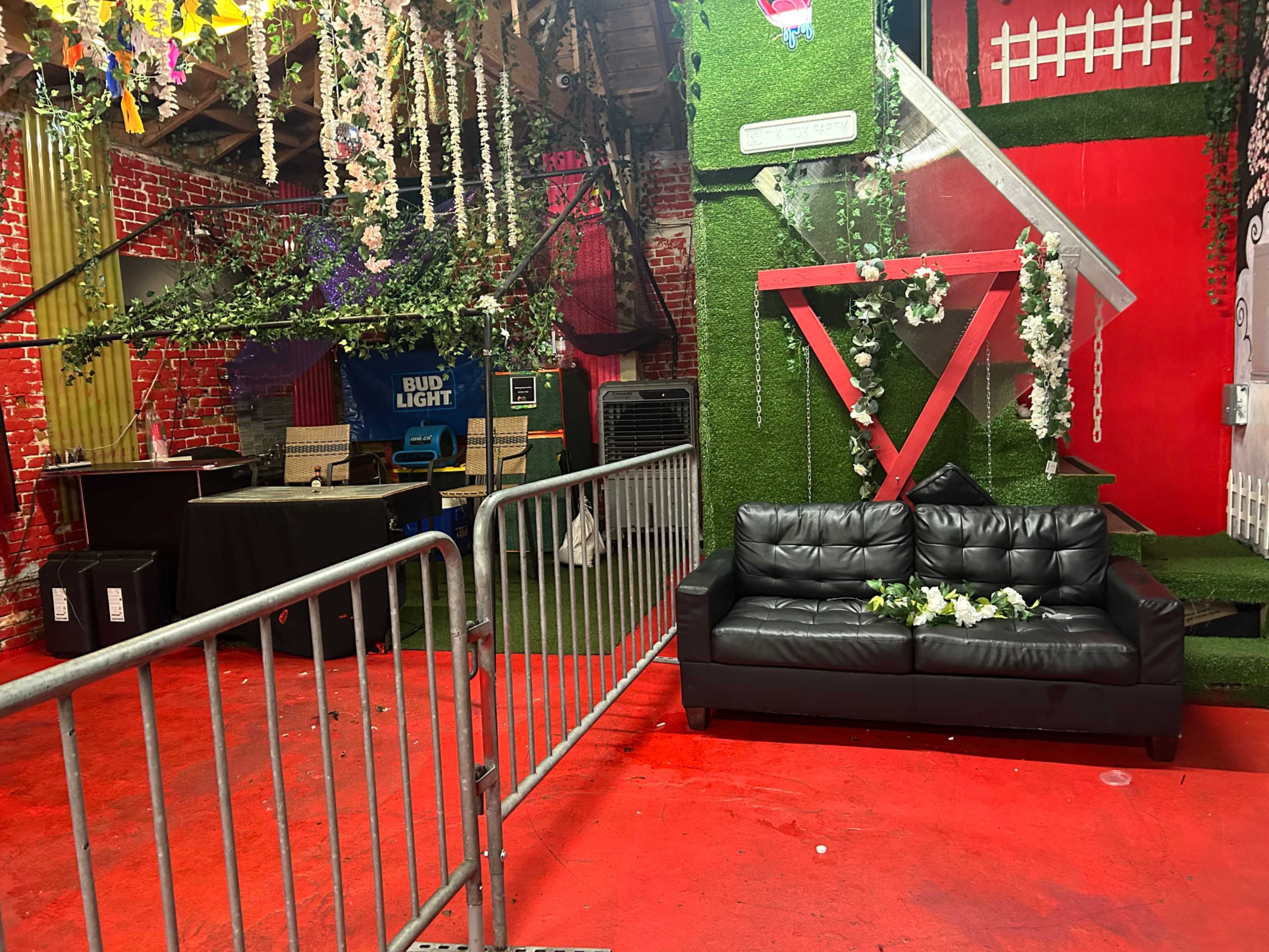 The image shows an indoor space decorated with greenery and floral arrangements, featuring a black couch and barriers separating different areas.