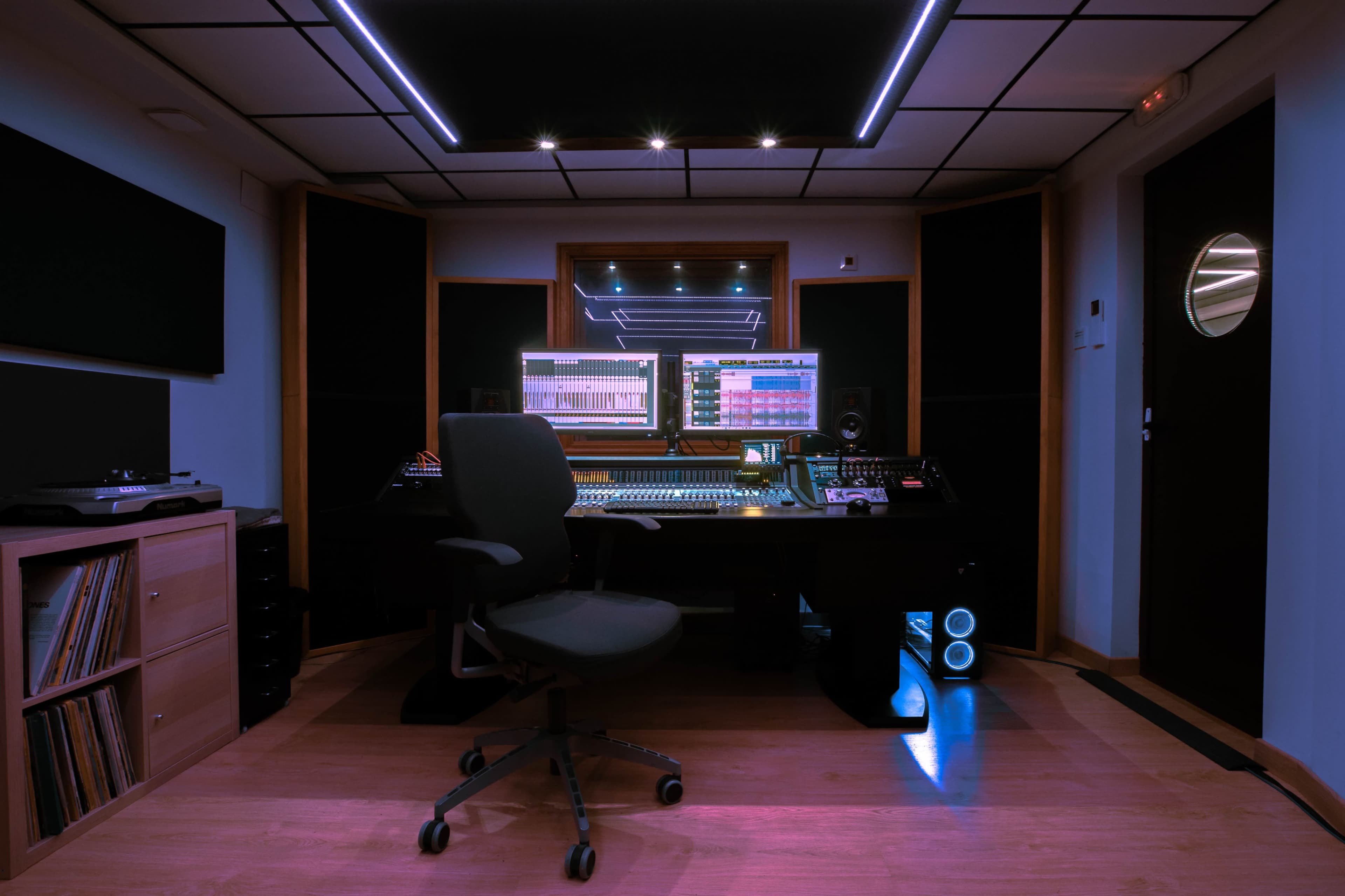 A recording studio features a mixing console, computer screens displaying audio tracks, and soundproof walls, with a swivel chair positioned in front of the control panel.