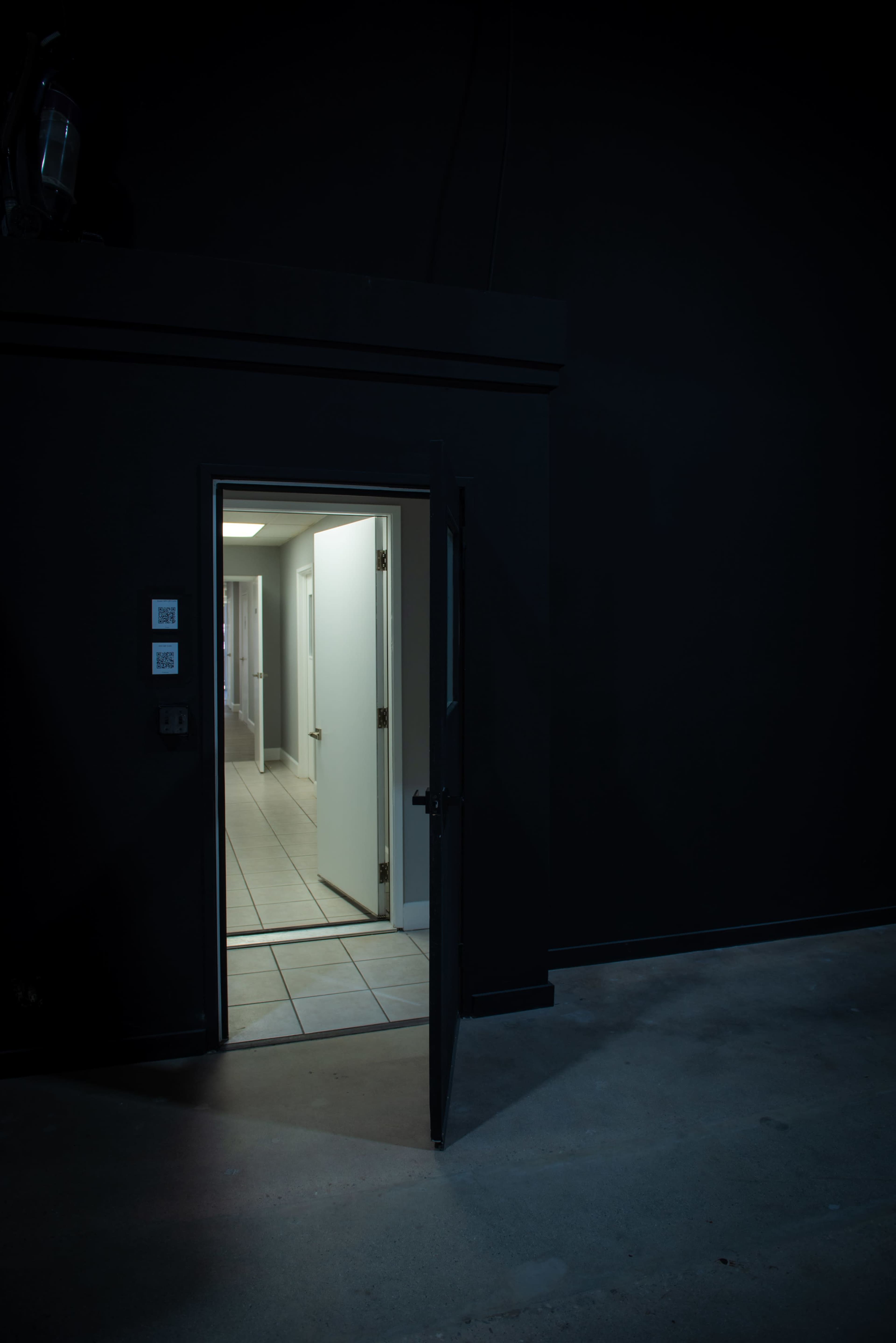 An open door reveals a brightly lit hallway with white walls and tiled flooring, contrasting against the dark surroundings.