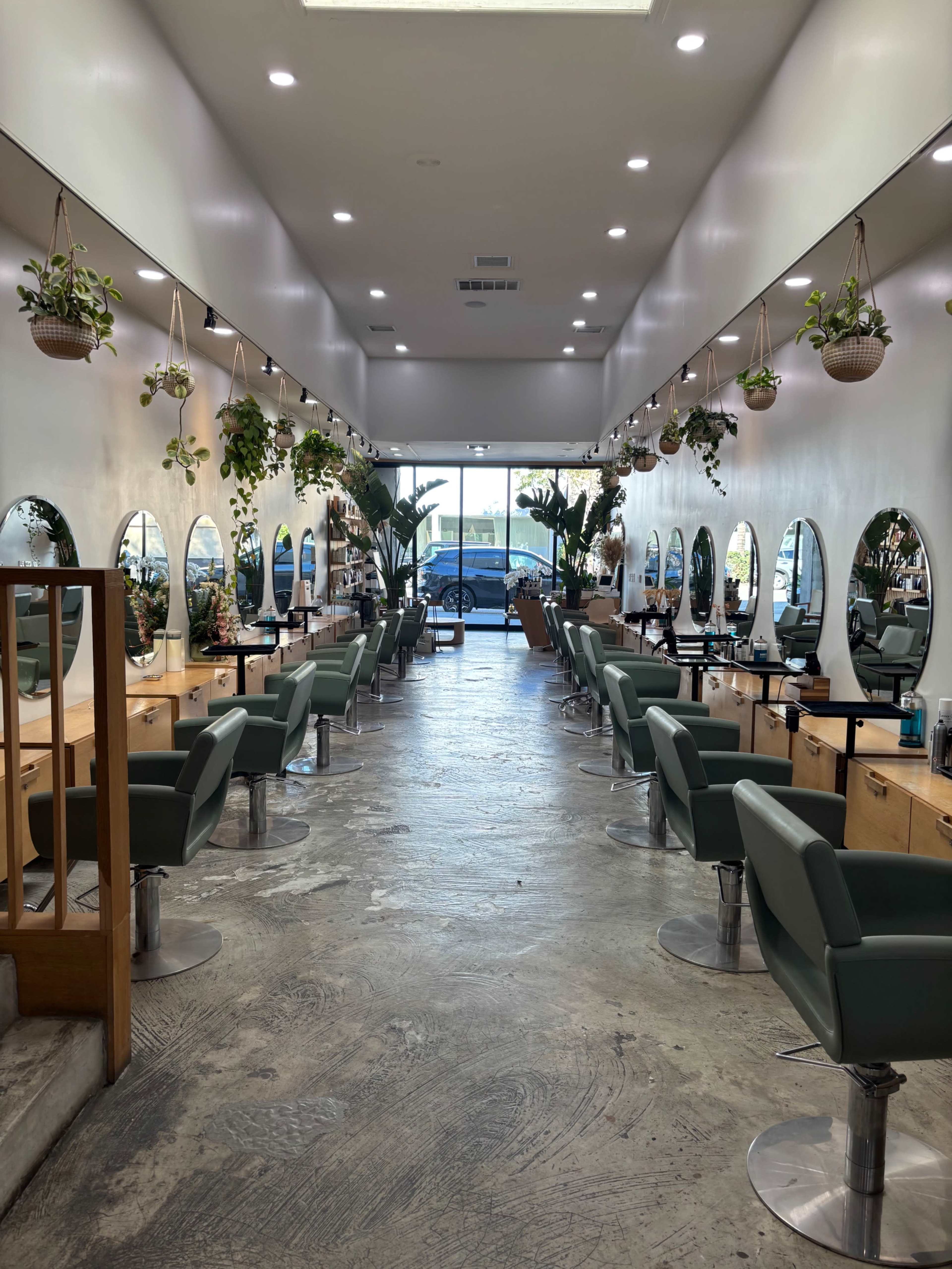 The image shows the interior of a modern hair salon with several styling stations lined along a spacious corridor, adorned with plants and large mirrors.