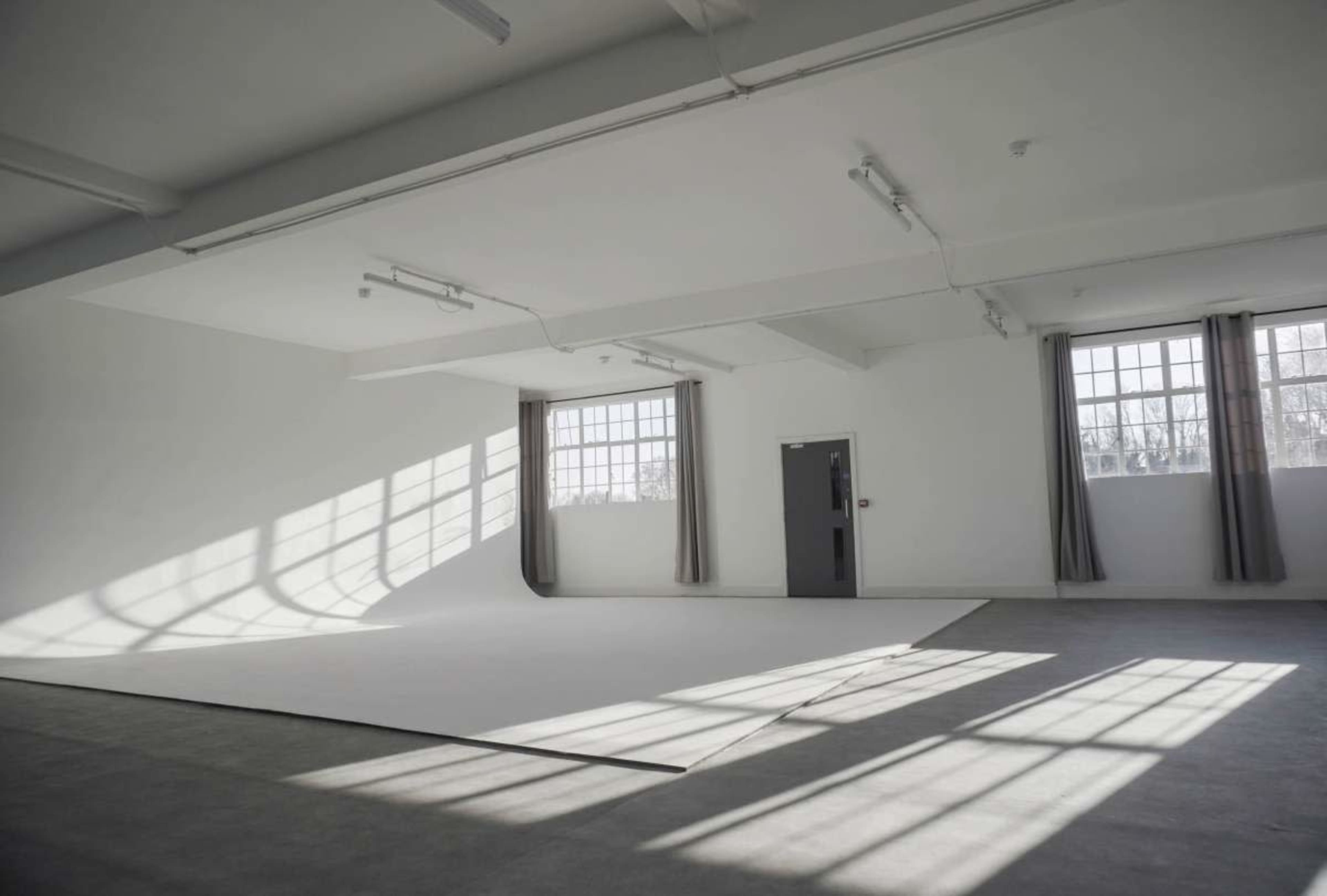 The image shows a spacious, empty room with white walls, large windows, and shadows cast on the floor.