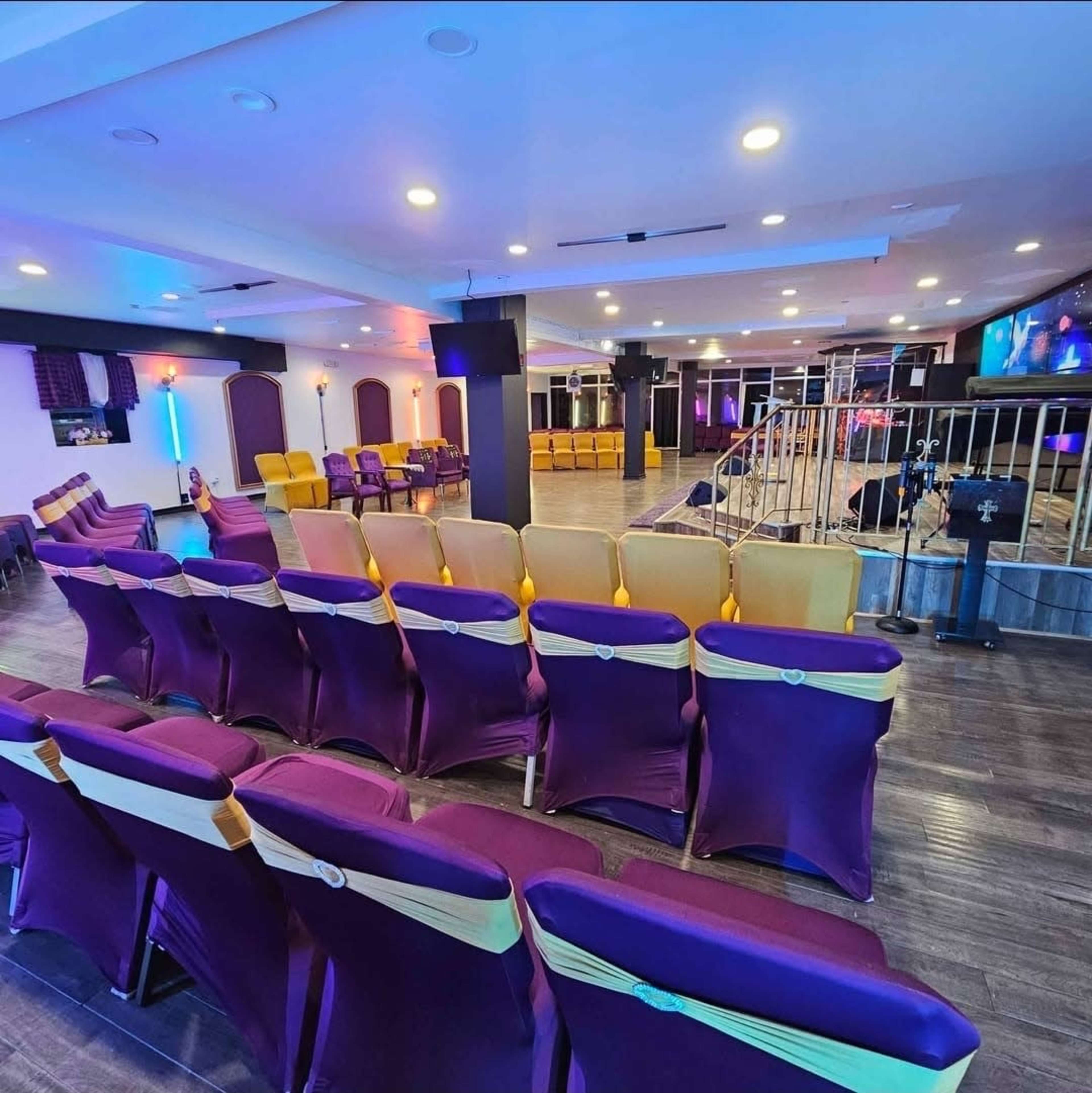The image shows a spacious venue with rows of chairs covered in purple and gold fabric, arranged facing a small stage with lighting and a microphone setup.