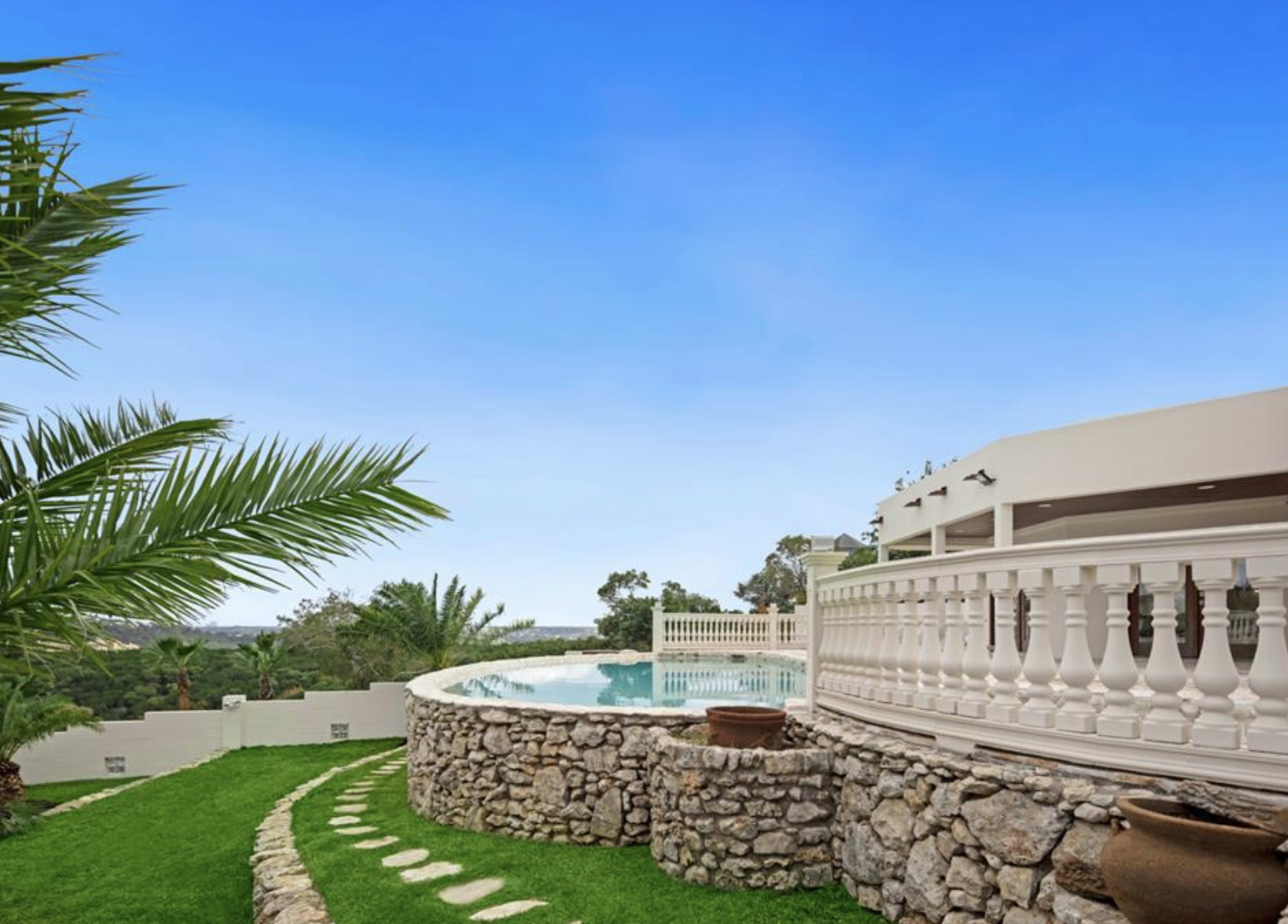 A stone wall surrounds a swimming pool with a view of greenery and a clear blue sky.