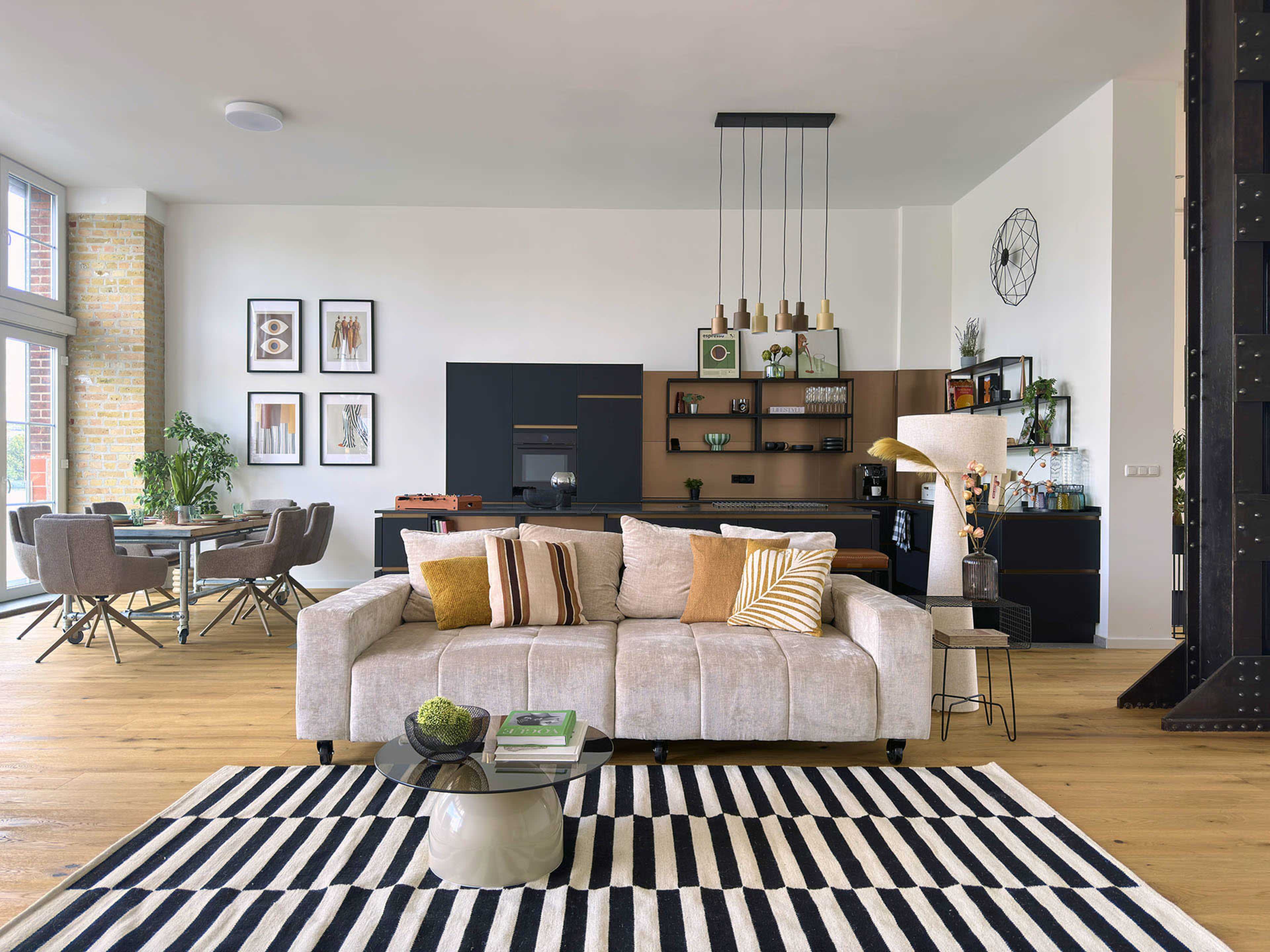 The image shows a modern living space featuring a plush sofa, a stylish dining area, and an open kitchen arrangement with contemporary decor.