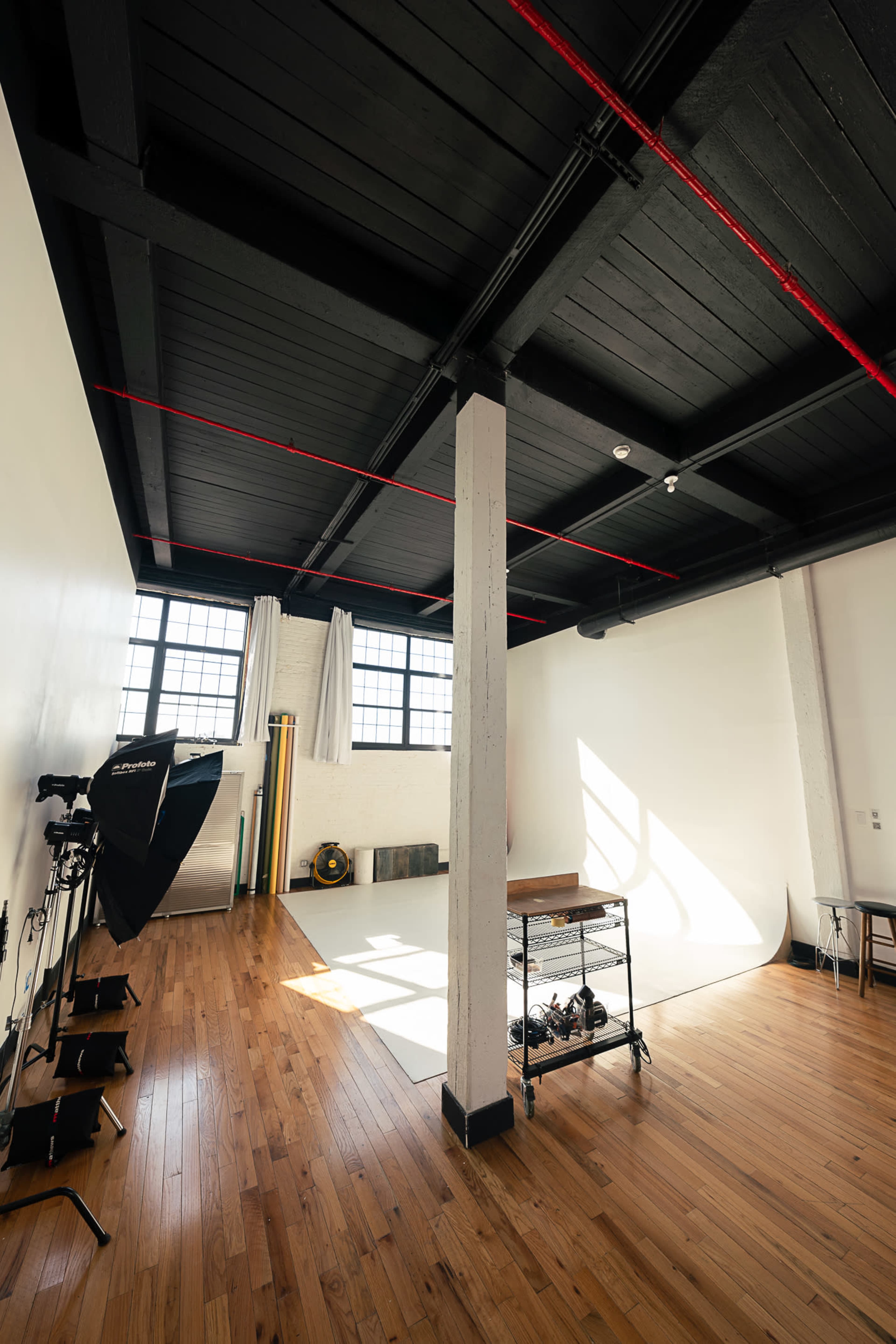 The image shows a spacious photography studio with a wooden floor, large windows, and various photography equipment arranged around a white backdrop.
