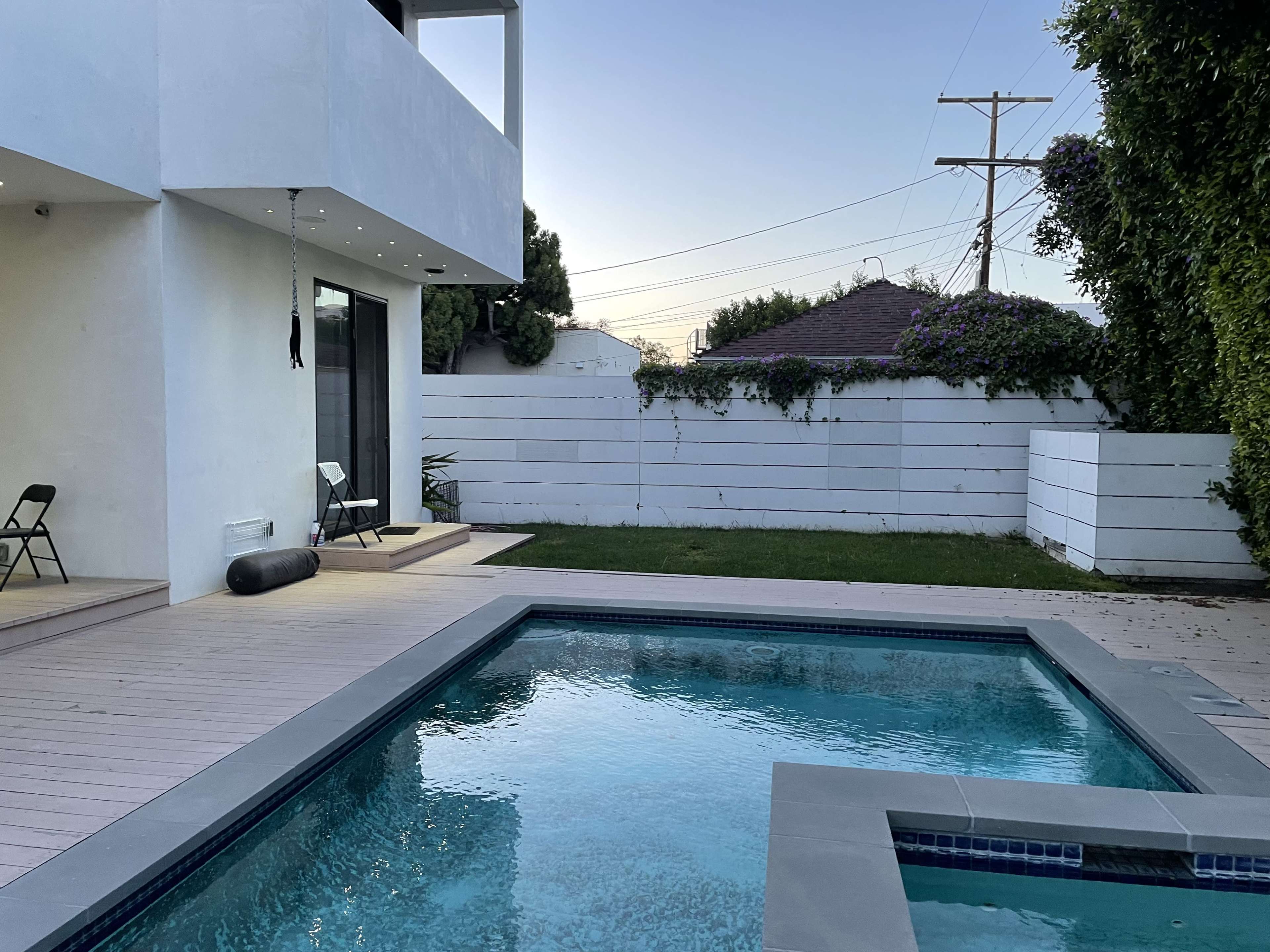 A modern backyard with a swimming pool, wooden deck, and a white fence surrounding a grassy area.