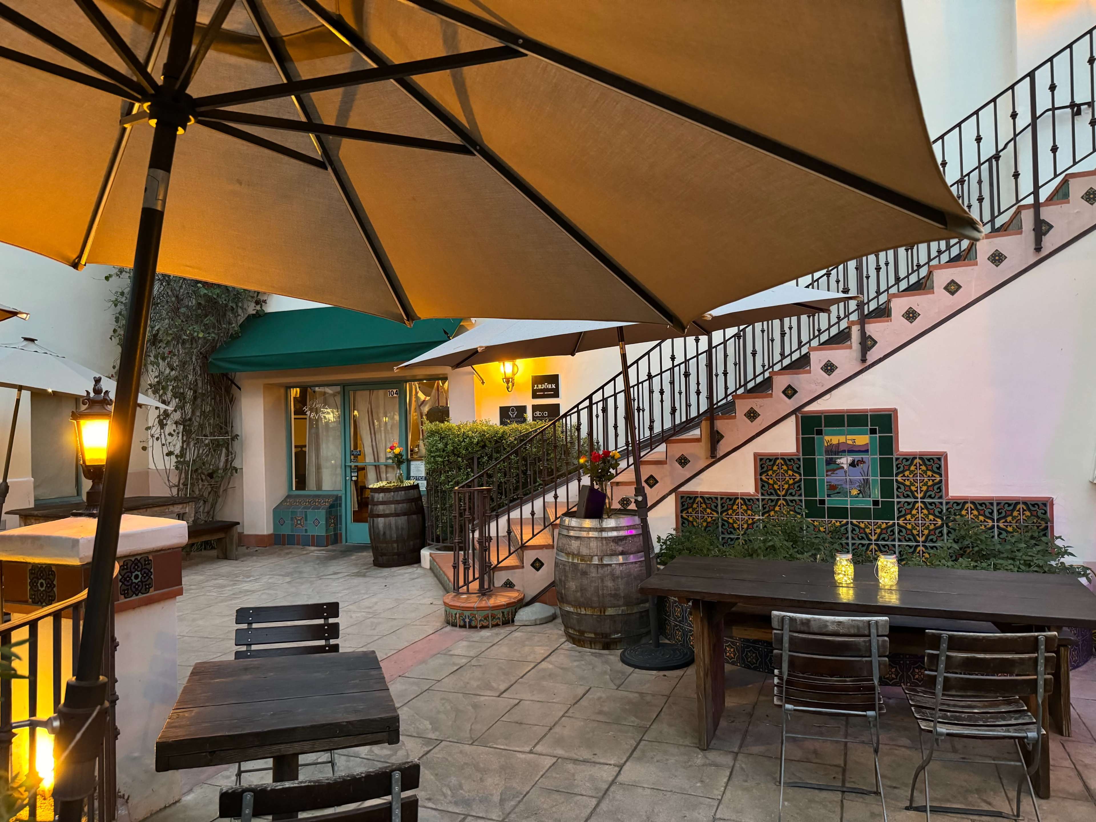 A cozy outdoor seating area with wooden tables, large umbrellas, and a staircase leading to a second level, surrounded by vibrant tile designs and greenery.