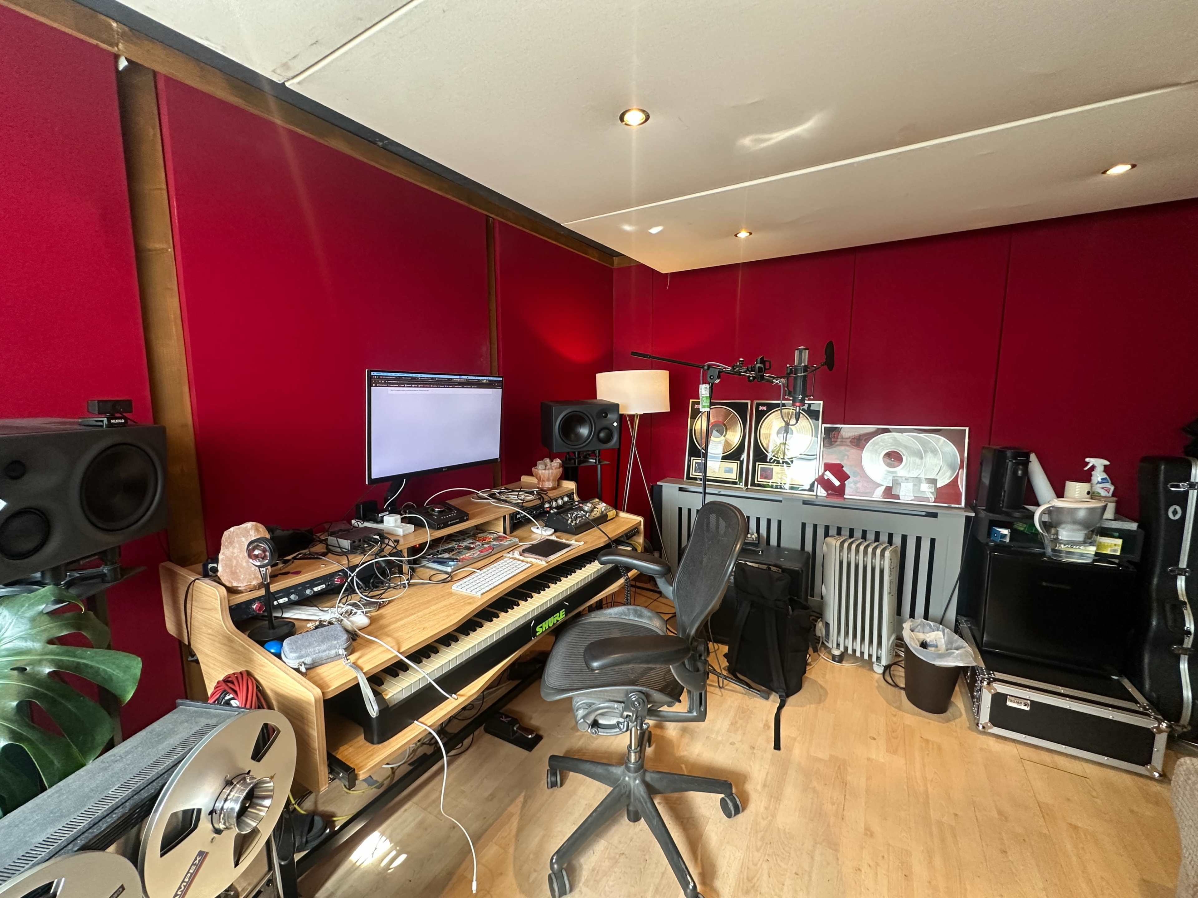 The image shows a music studio featuring a keyboard, a computer monitor, studio monitors, and various electronic equipment against red walls.