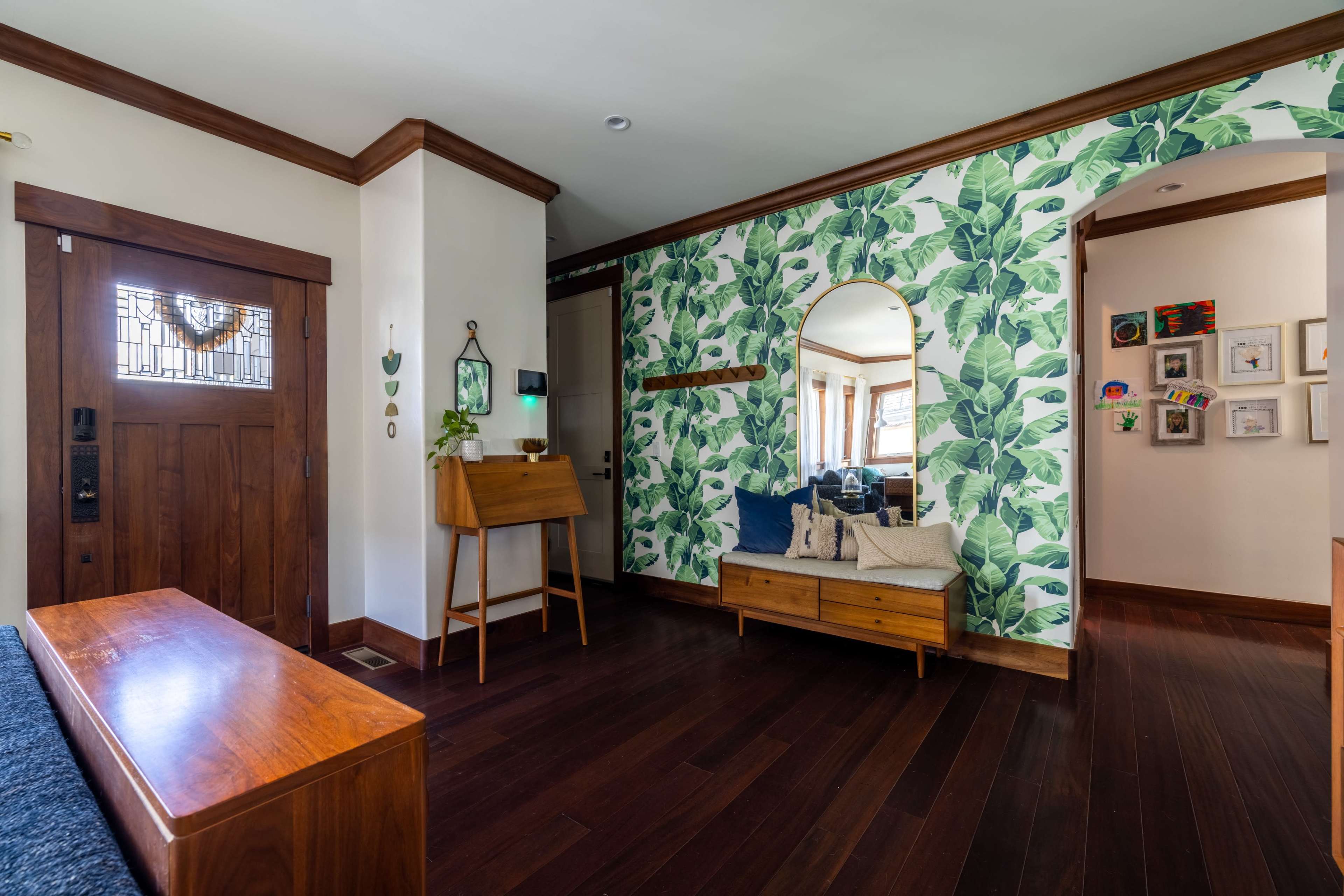 A living room with a wooden entrance door, a leaf-patterned feature wall, a wooden bench, and framed pictures displayed on the wall.