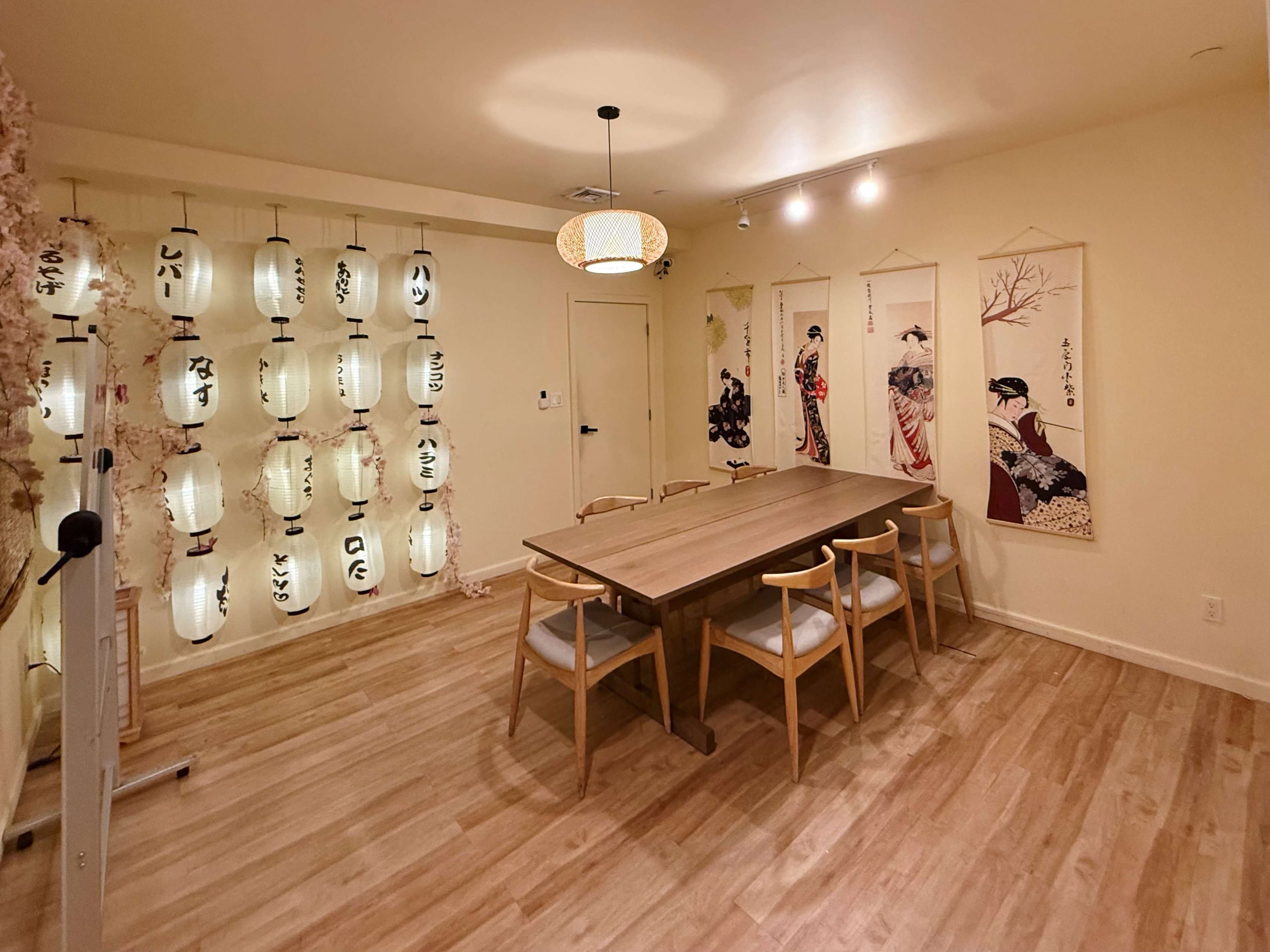 The image shows a dining room with a large wooden table, surrounded by chairs, and decorated with hanging lanterns and wall art featuring historical Japanese figures.