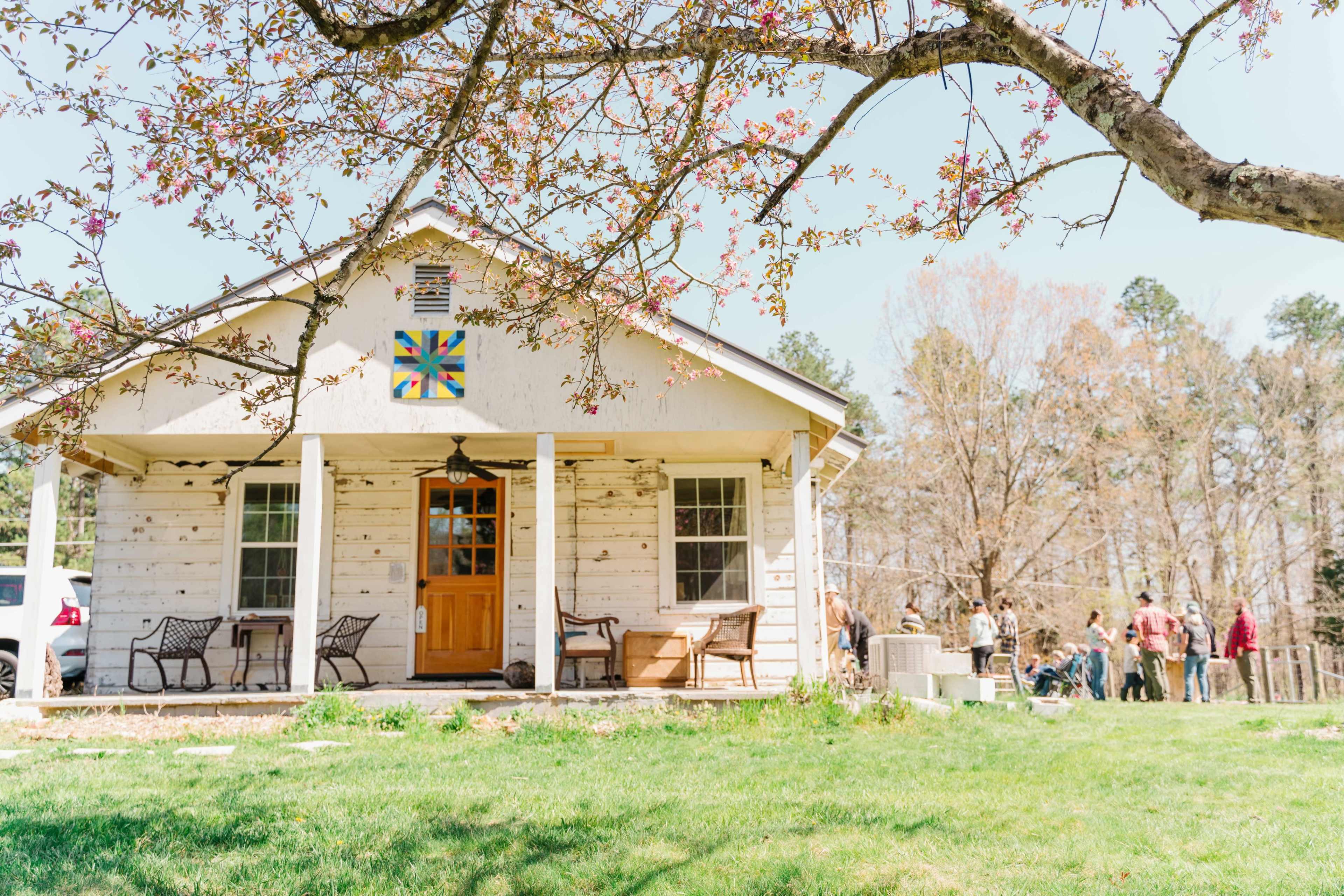 A rustic house with a flower-patterned quilt square on the front, surrounded by a grassy yard and several people gathered nearby.