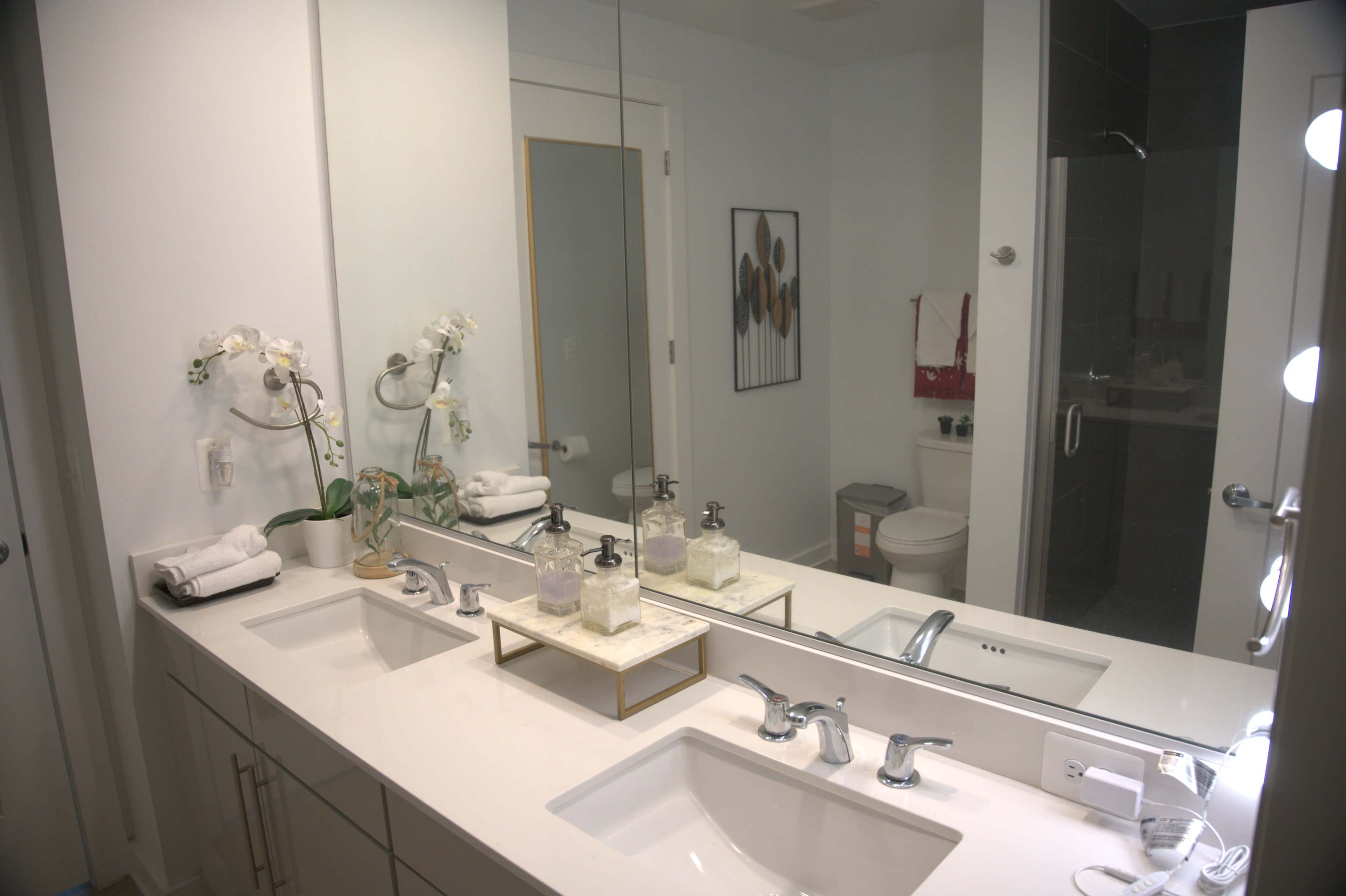 A modern bathroom with a double sink vanity, a large mirror, and a shower area visible in the background.