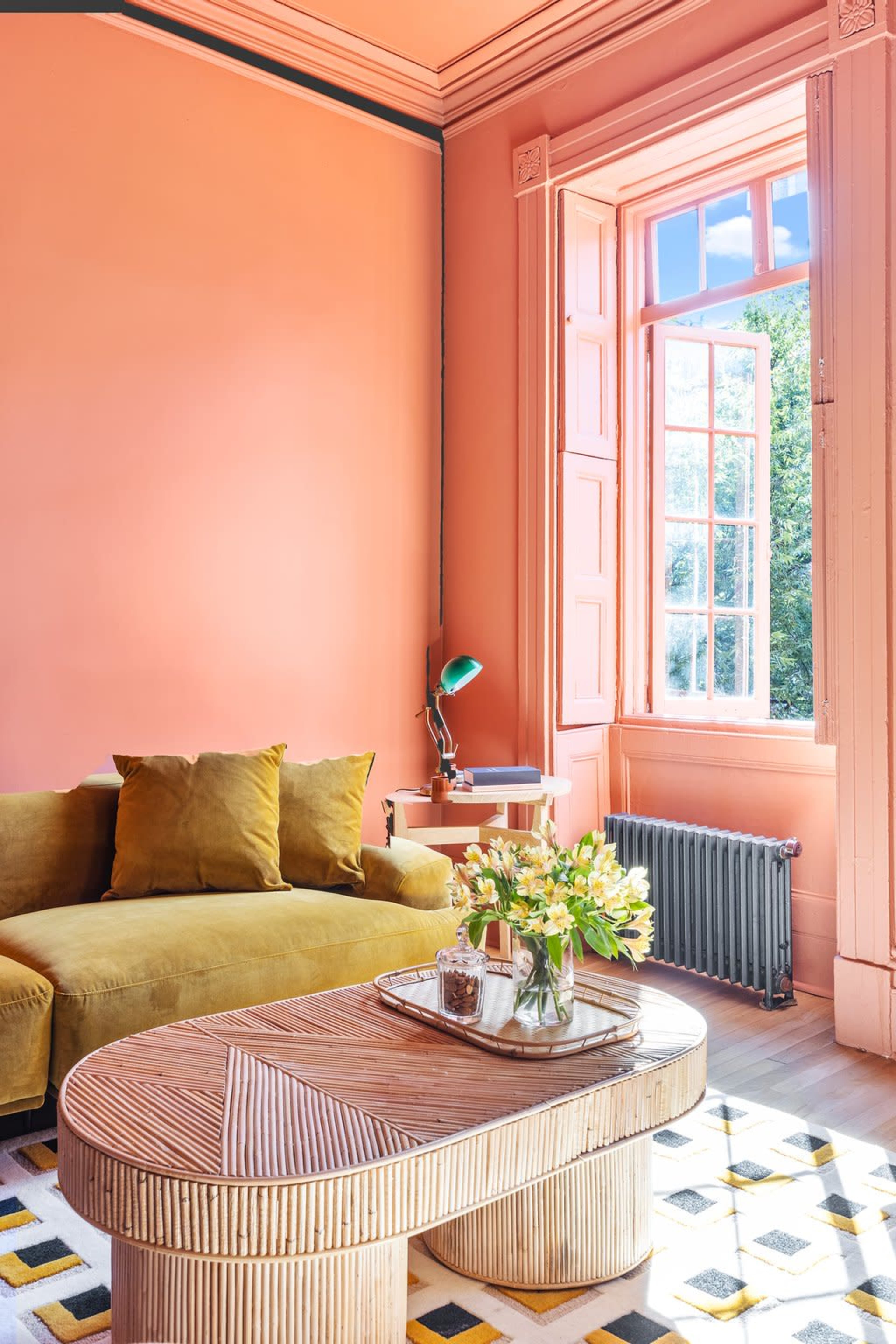 A bright living room features a green sofa with pillows, a wooden coffee table, and a large window with colorful trim that lets in natural light.