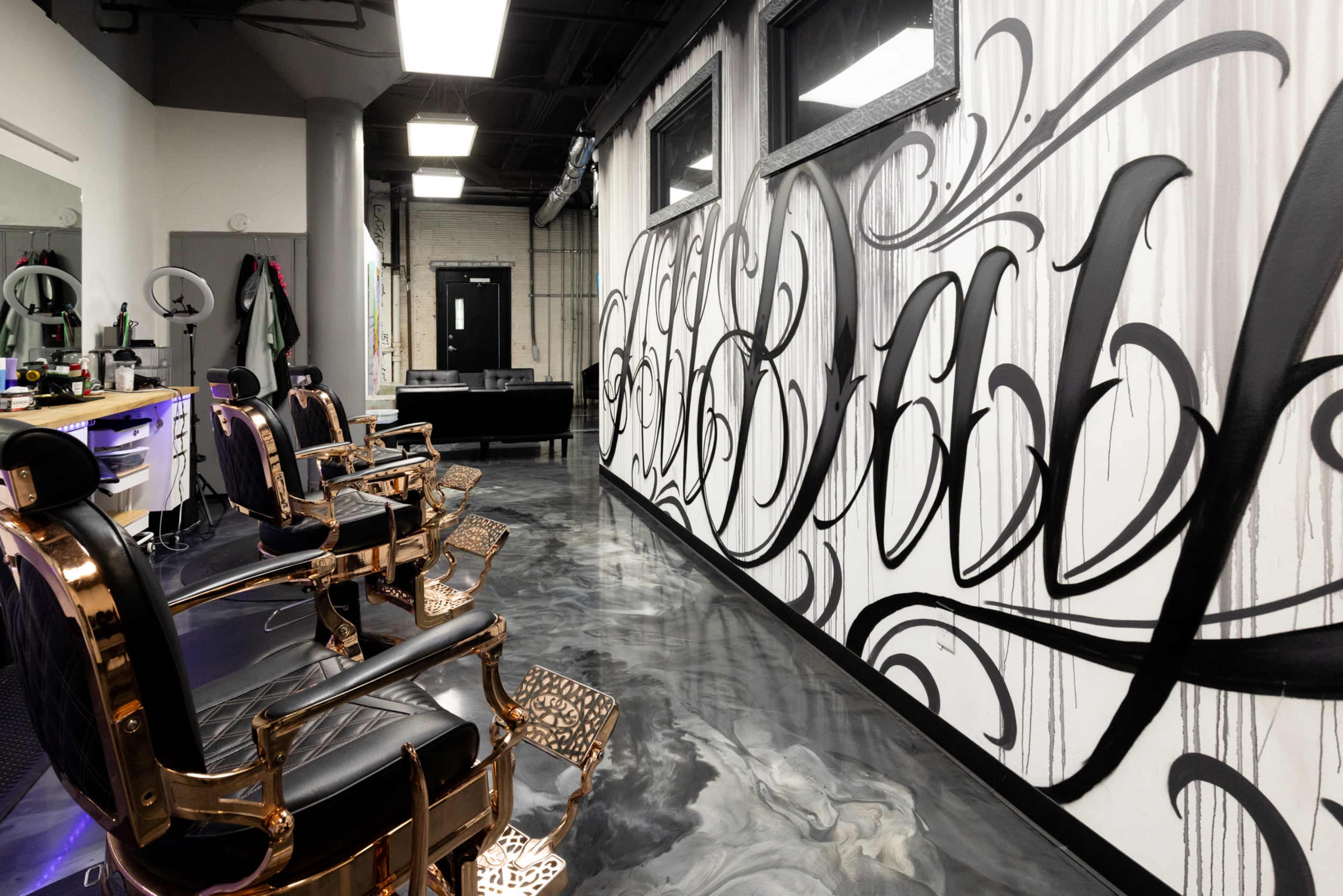 The image depicts a stylish barbershop interior featuring two ornate barber chairs facing a wall adorned with large, artistic calligraphy.