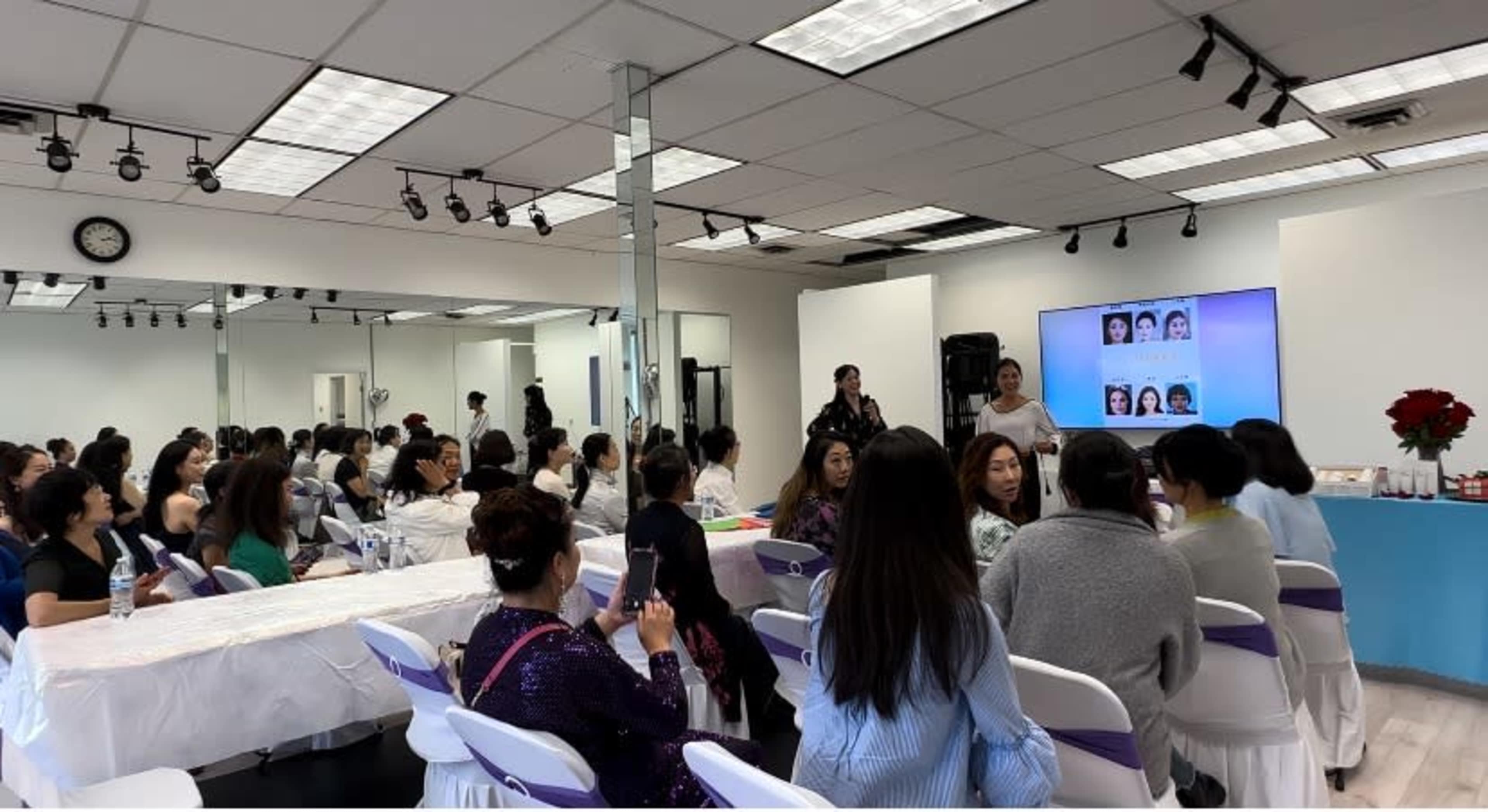 A large group of people attentively listens to a presentation in a well-lit seminar room with rows of chairs and a screen displaying images.