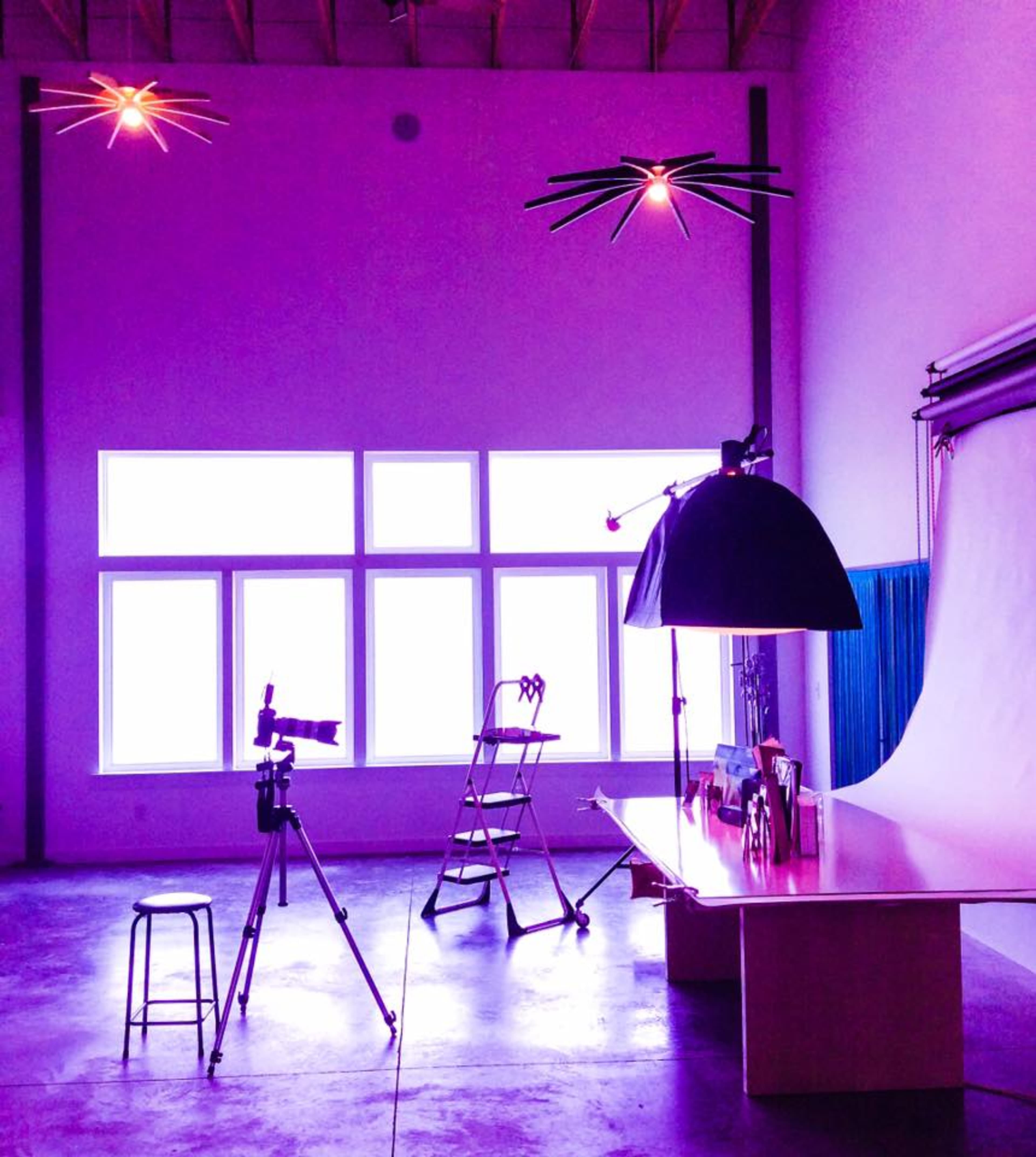 A photography studio features a large backdrop, a stool, a camera on a tripod, and a lighting setup under purple-hued ceiling lights.