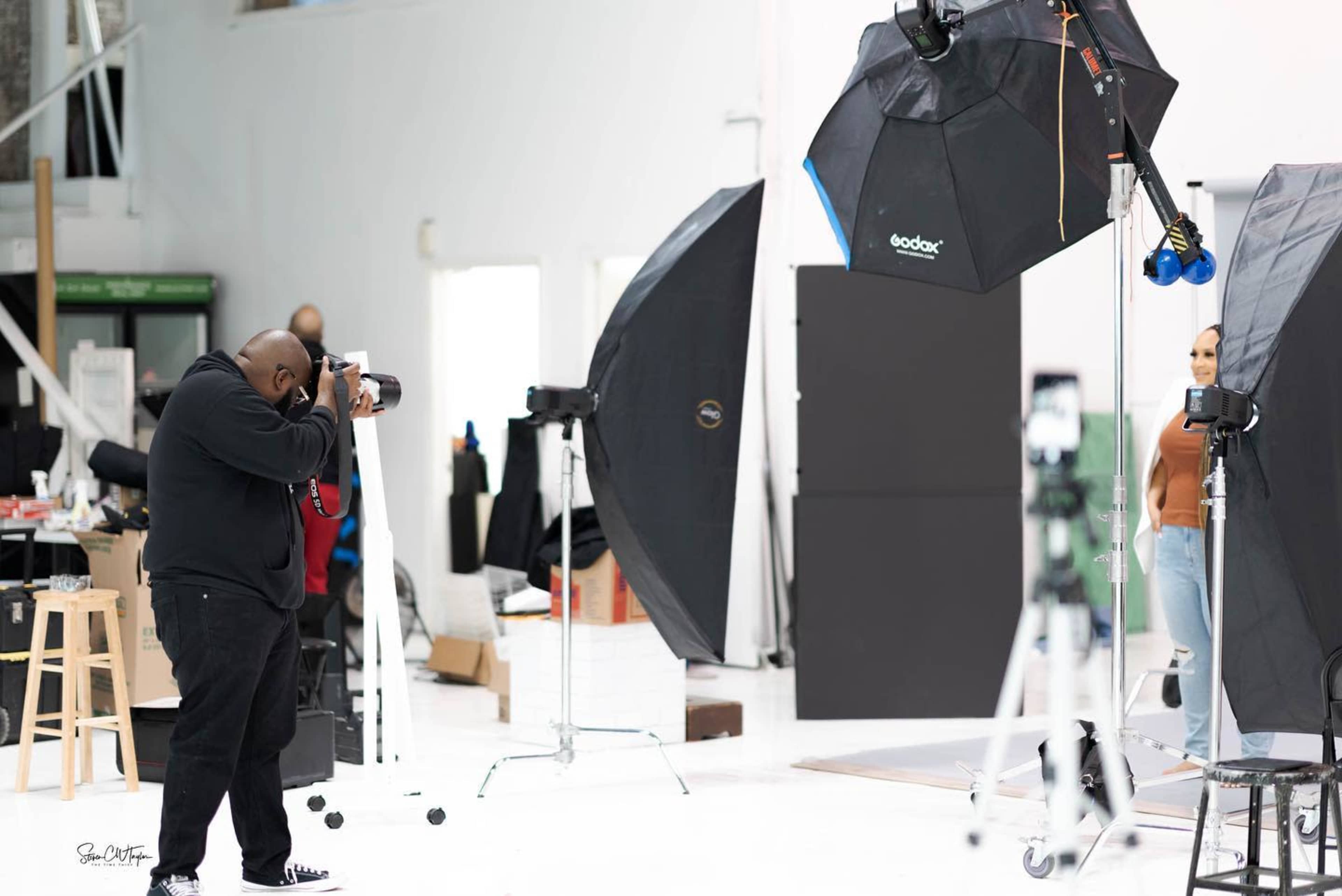 A photographer takes pictures of a model in a brightly lit studio, surrounded by softboxes and backdrop stands.