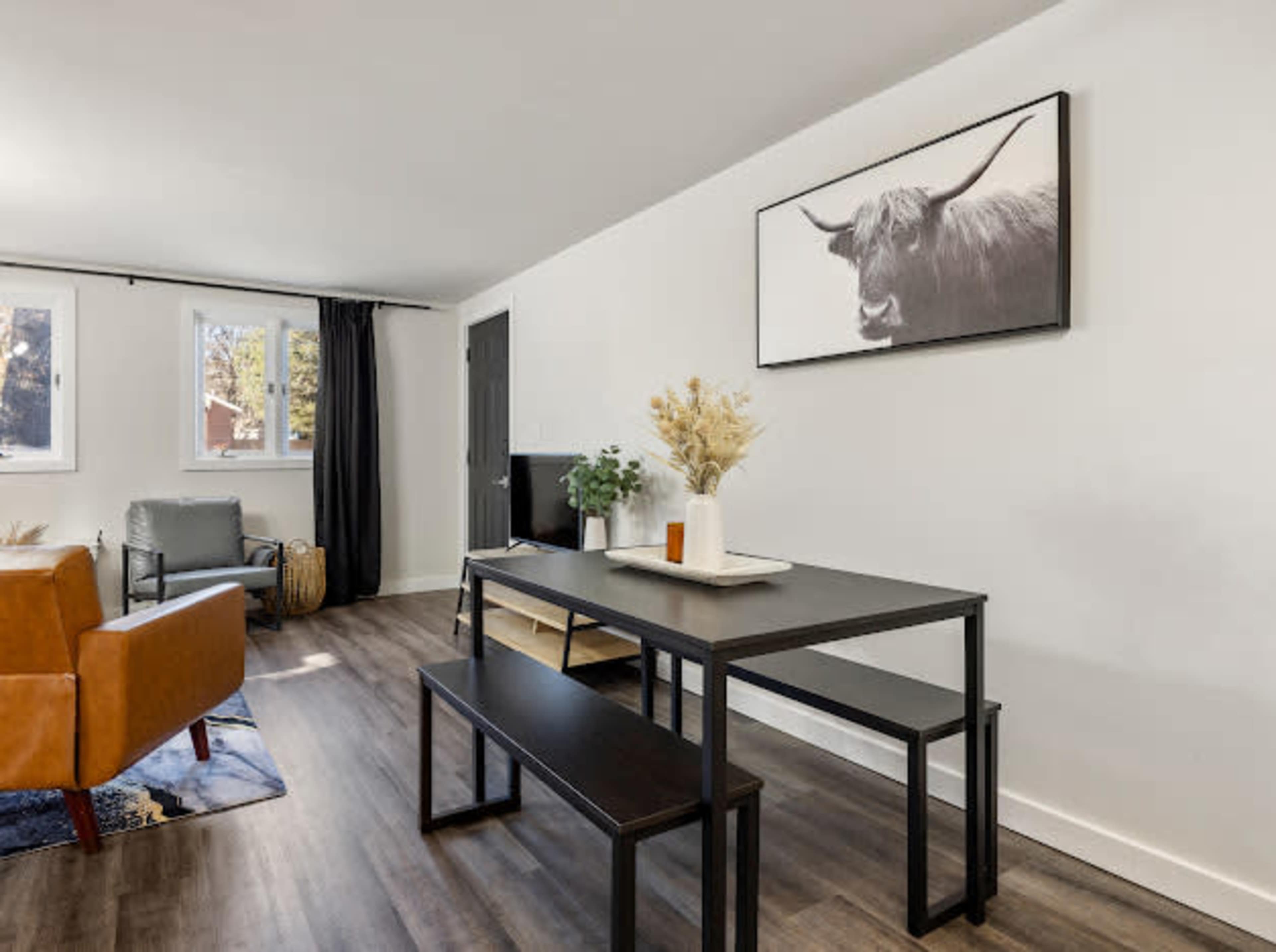 A modern living space featuring a dining table with benches, a brown sofa, a TV on a stand, and a large wall art piece of a cow.