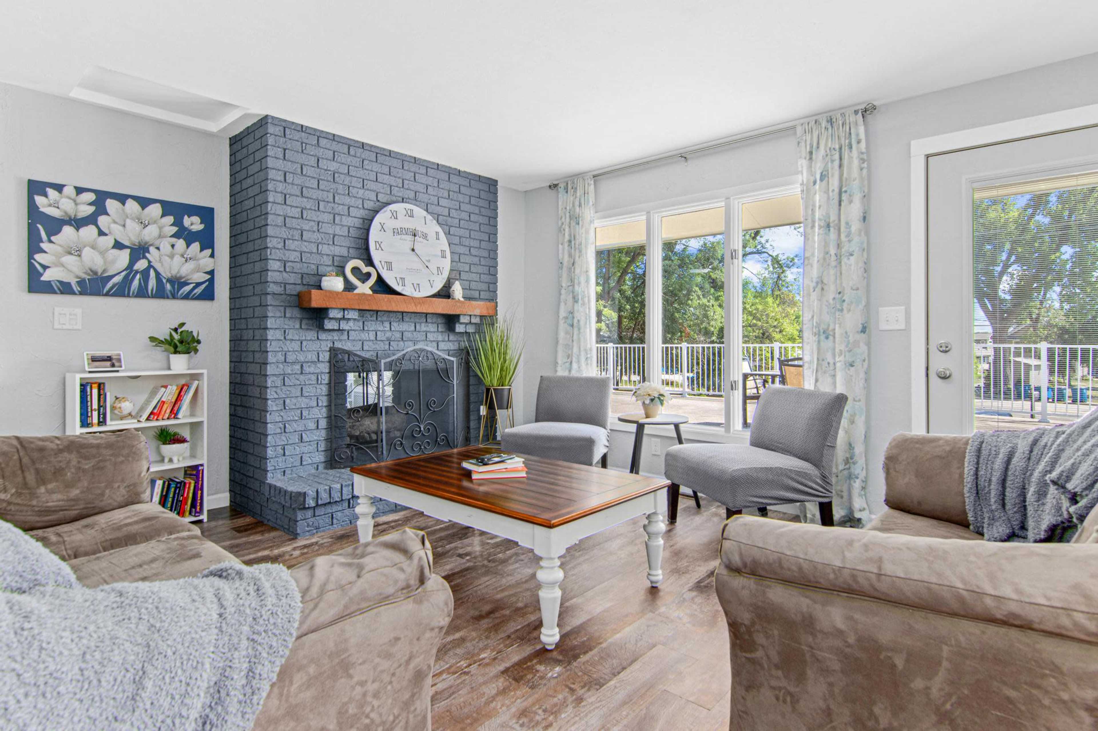 A cozy living room featuring a blue brick fireplace, a wooden coffee table, and two gray chairs, with large windows overlooking an outdoor area.