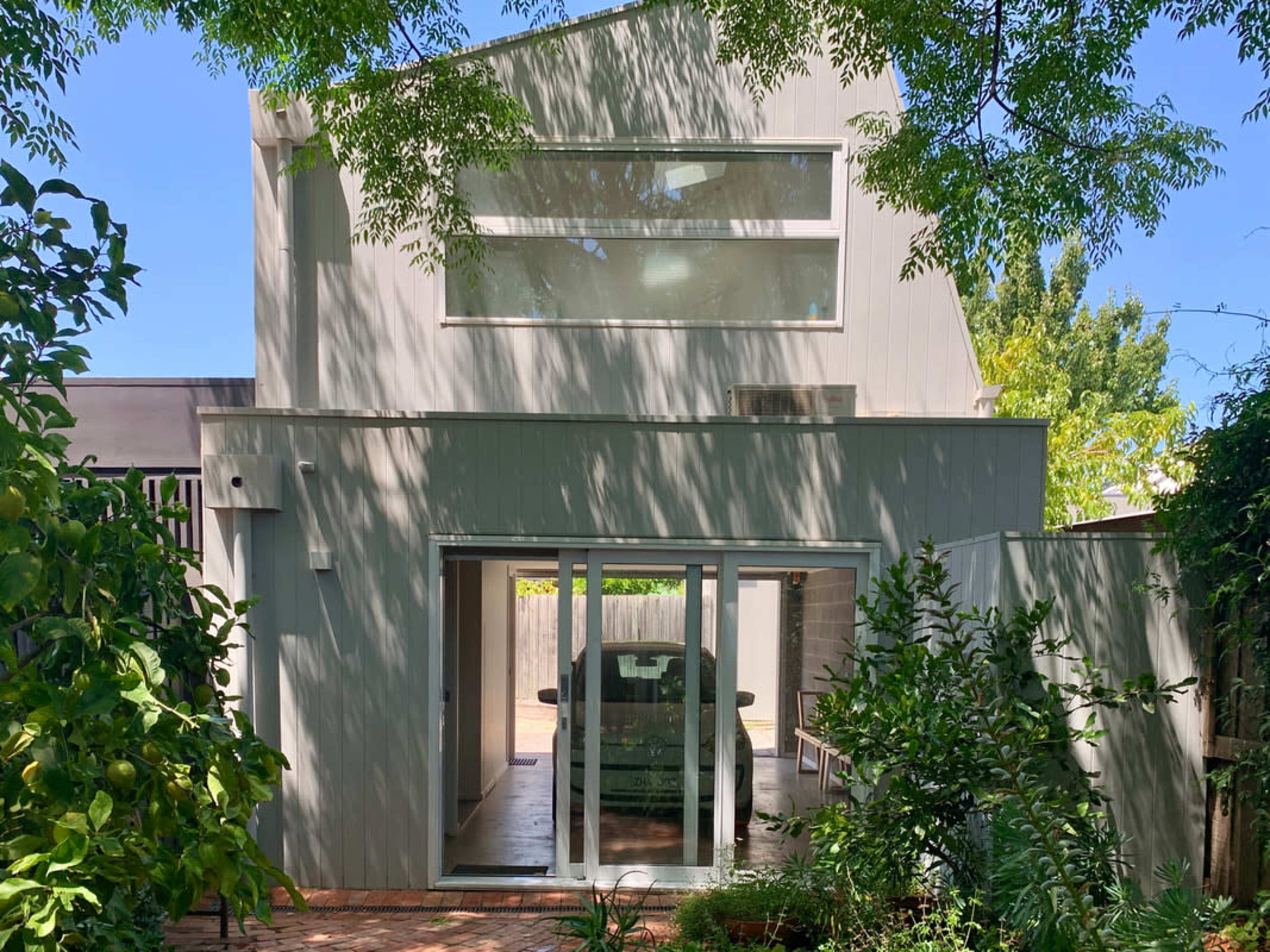A two-story house with large windows and a car visible through the sliding glass doors is surrounded by greenery.