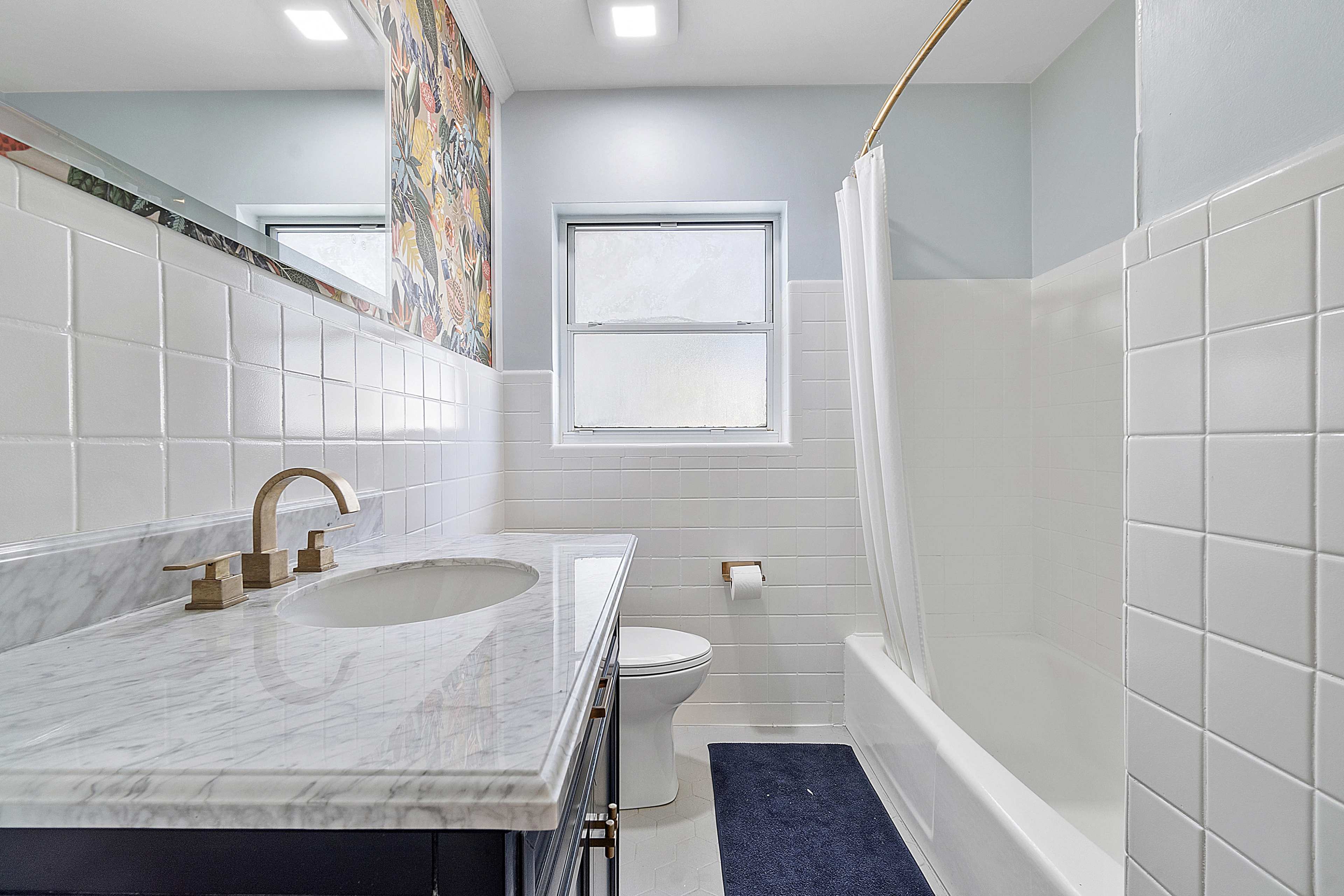 A bathroom featuring a marble countertop, a white tiled bathtub with a shower curtain, and a window allowing natural light.