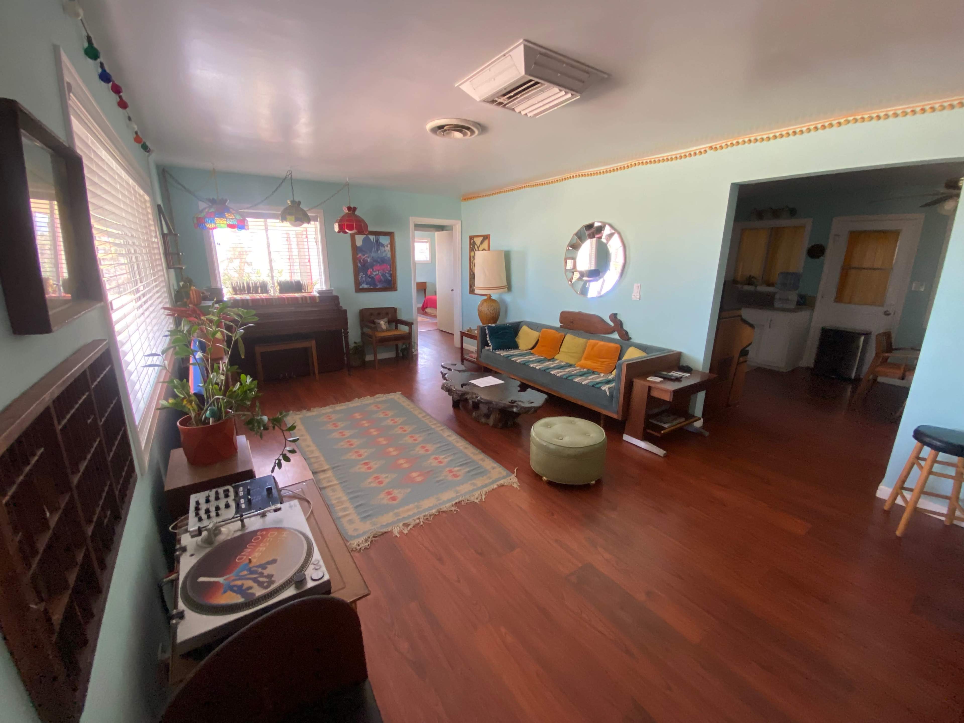 The image shows a bright living space with a wooden floor, a record player, a sofa, and a dining area in the background.