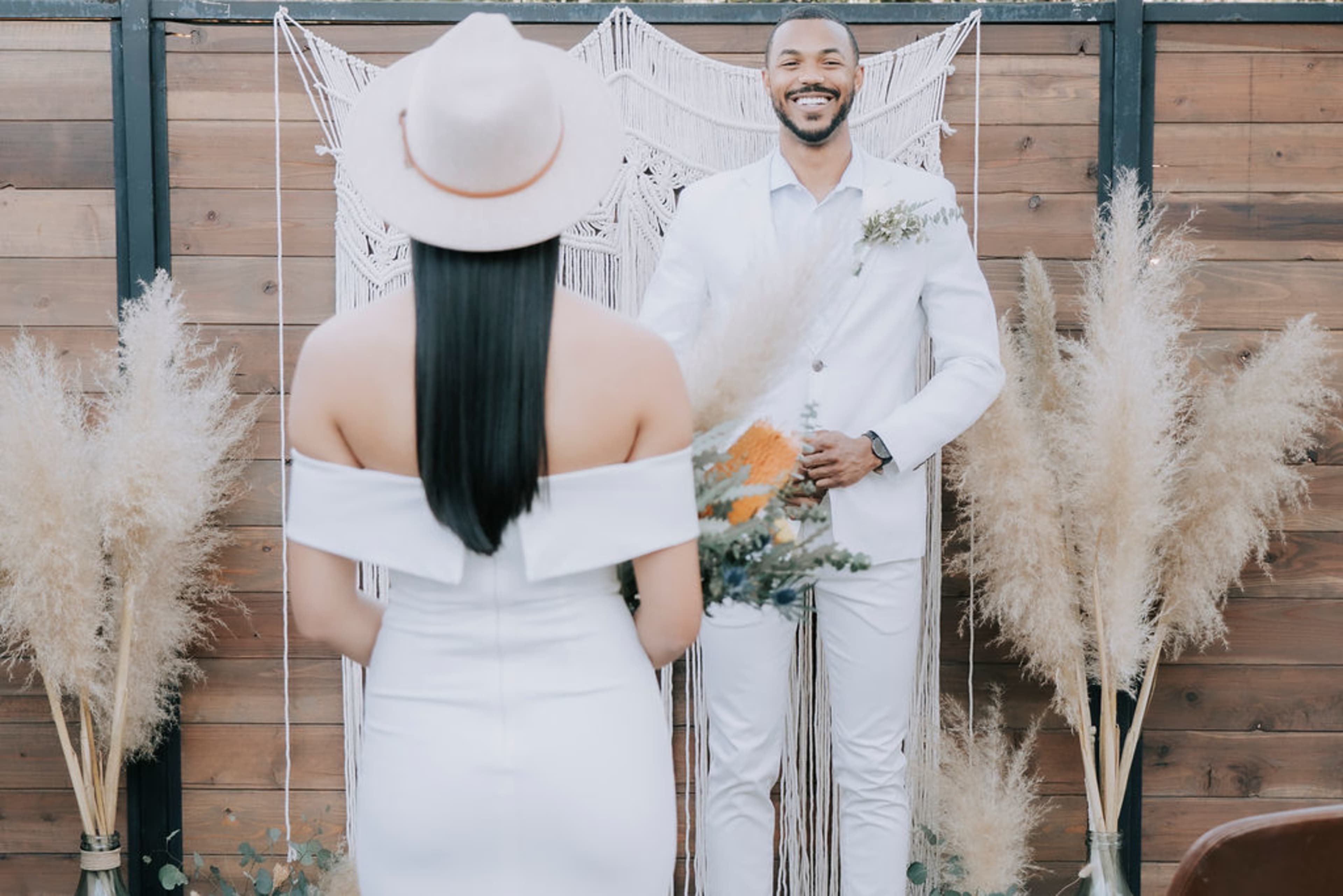 A couple dressed in white stands in front of a macramé backdrop adorned with pampas grass during a wedding ceremony.