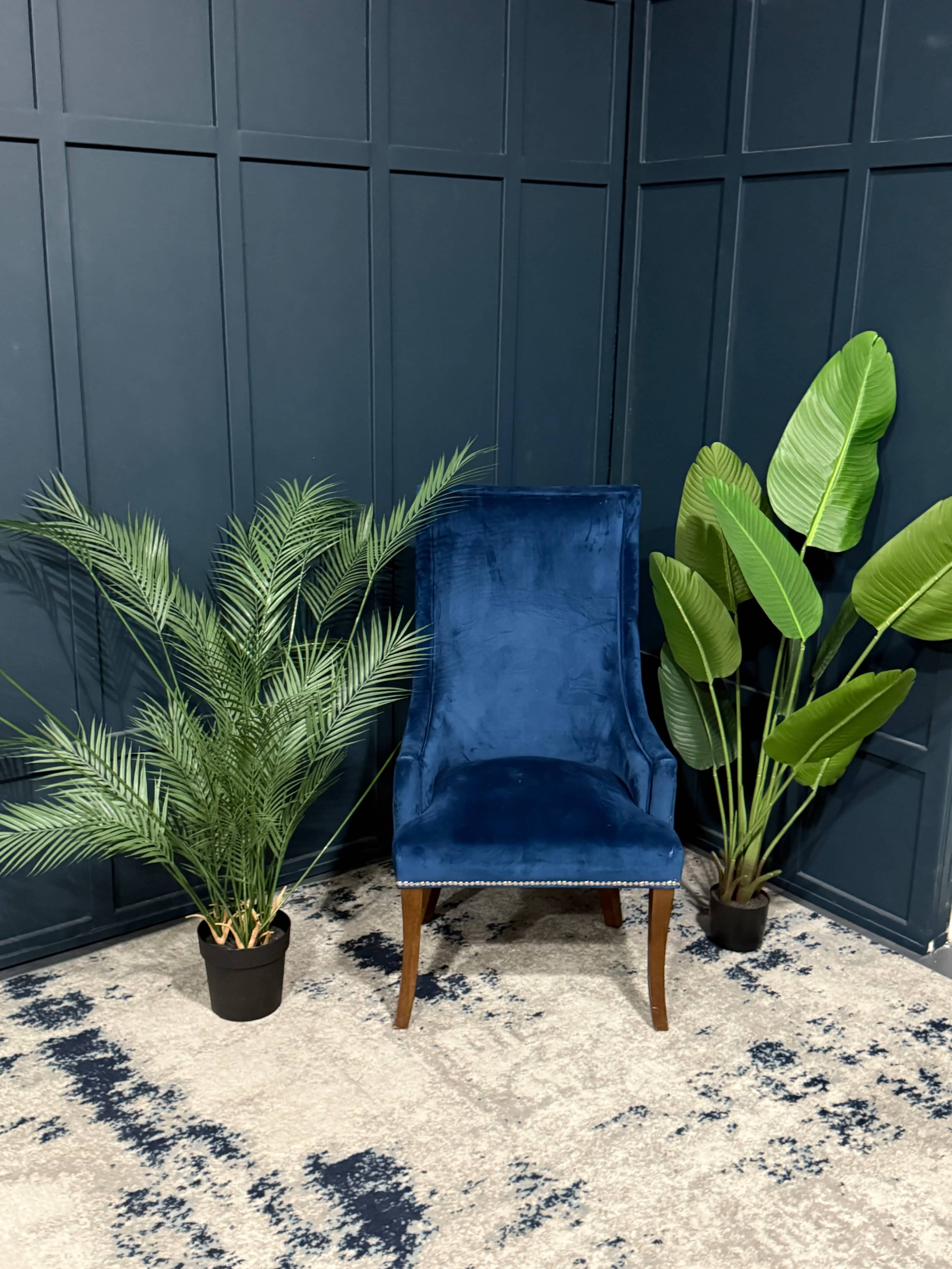 A velvet blue chair is positioned between two large green potted plants against a dark paneled wall.