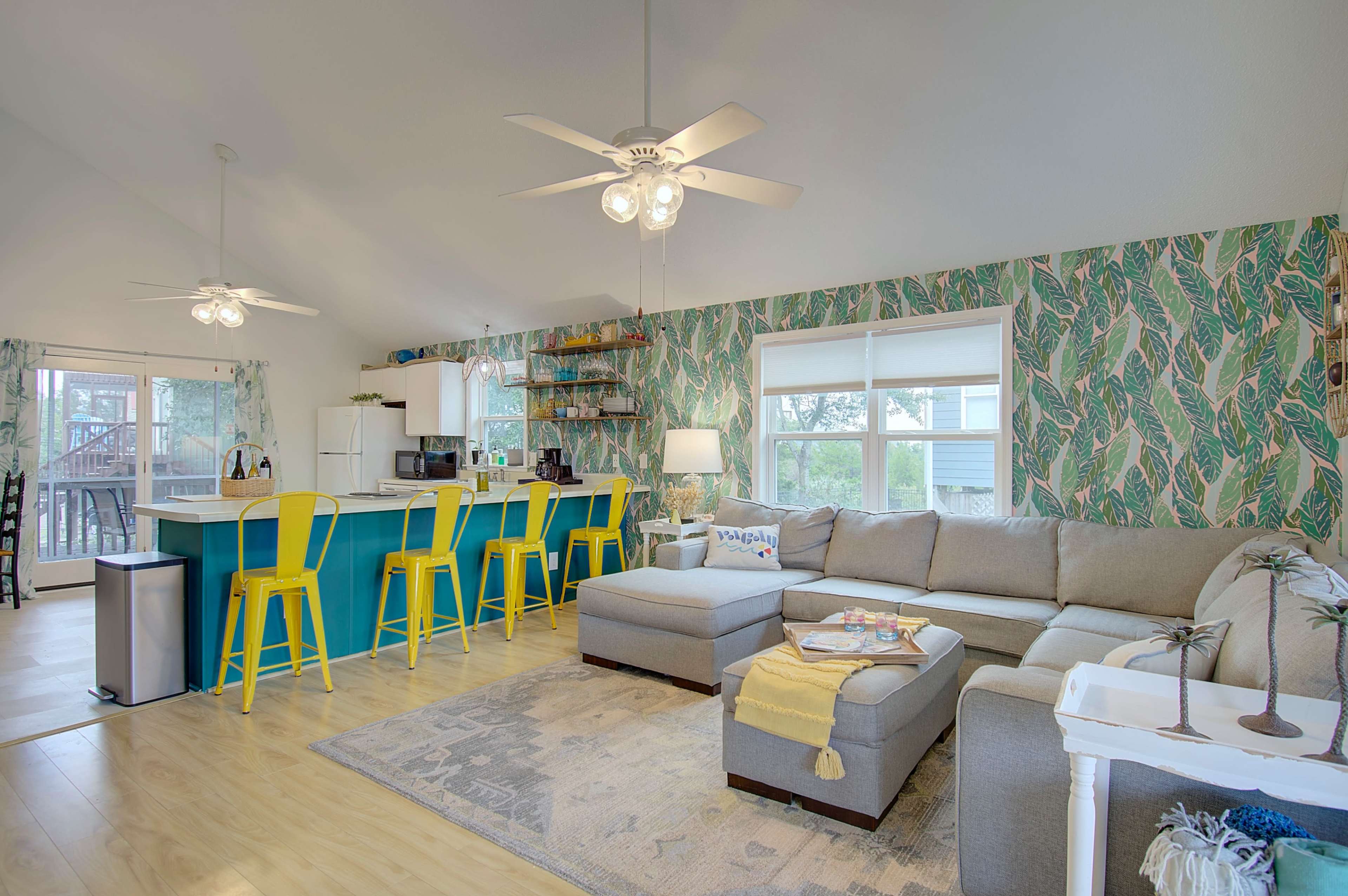 A bright, open living space with a sectional sofa, a kitchen island with yellow stools, and tropical-themed wallpaper.