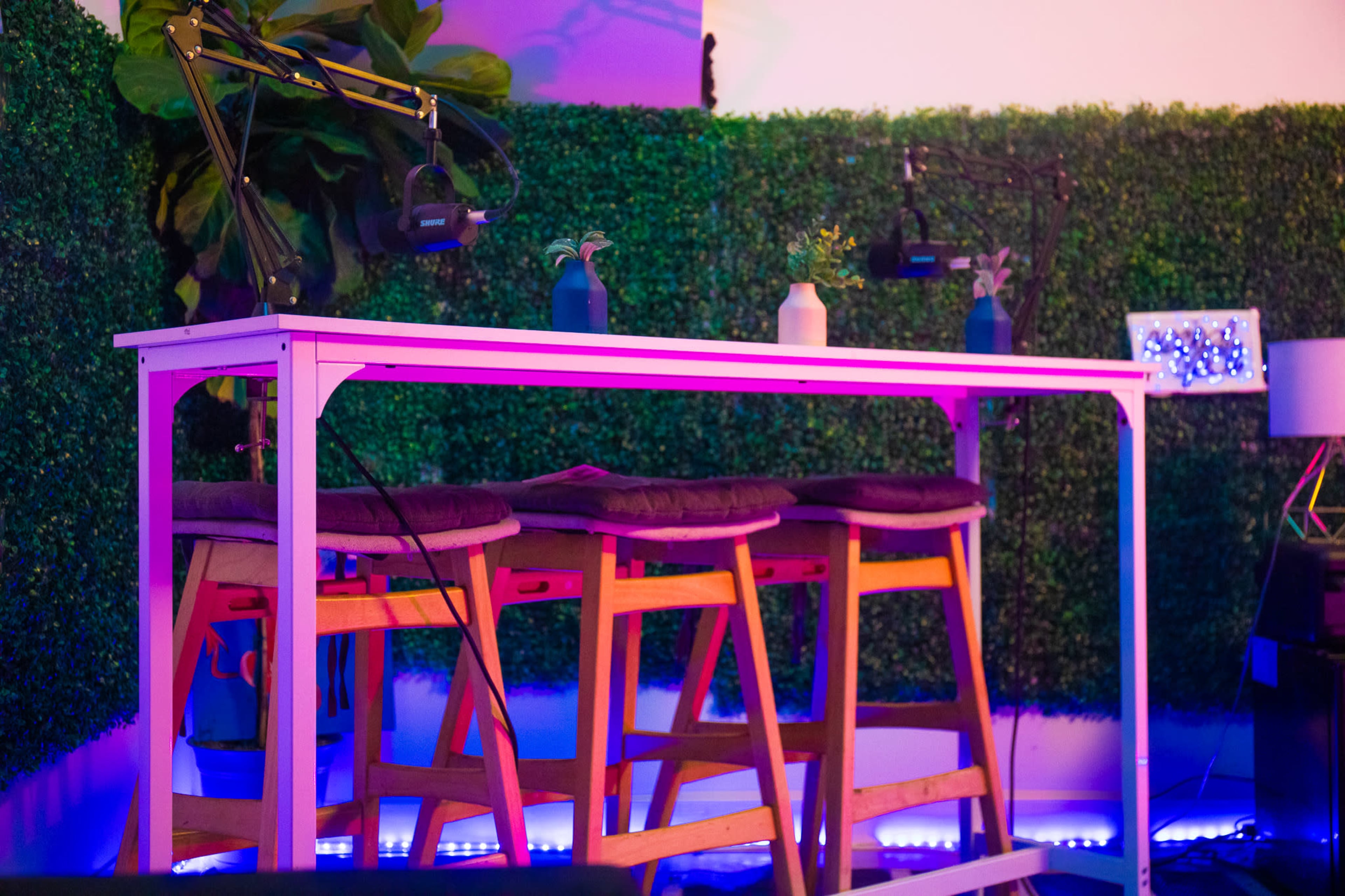 A modern, elevated table with three wooden stools is set against a backdrop of artificial greenery and colorful lighting.