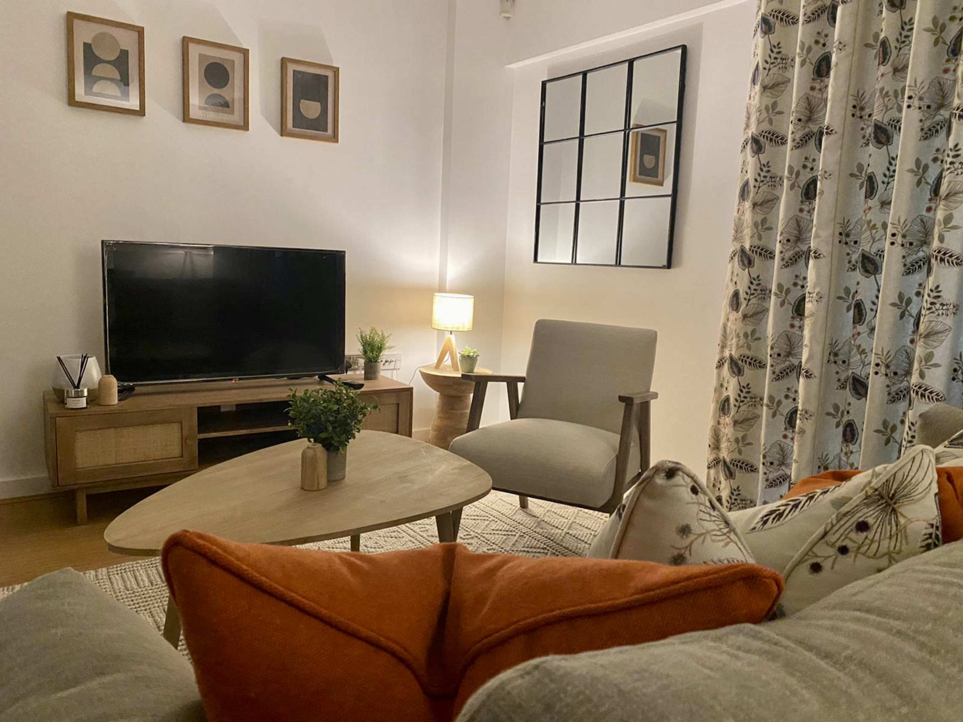 A cozy living room features a television, a wooden coffee table, an armchair, and decorative pillows on a sofa, with plants and artwork on the walls.