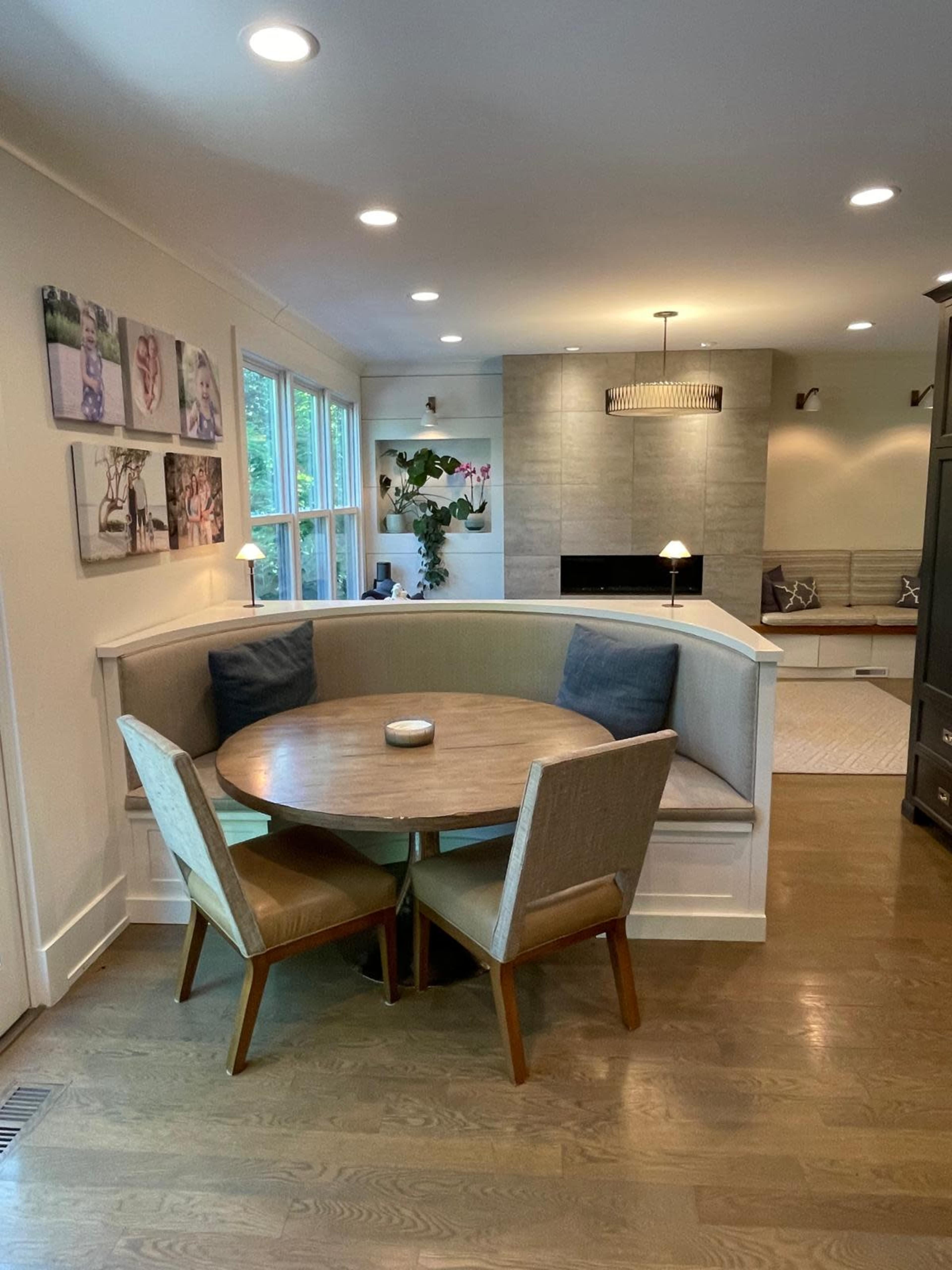 The image shows a cozy dining nook with a round table surrounded by a built-in bench and two chairs, set against a wall of windows and modern decor.