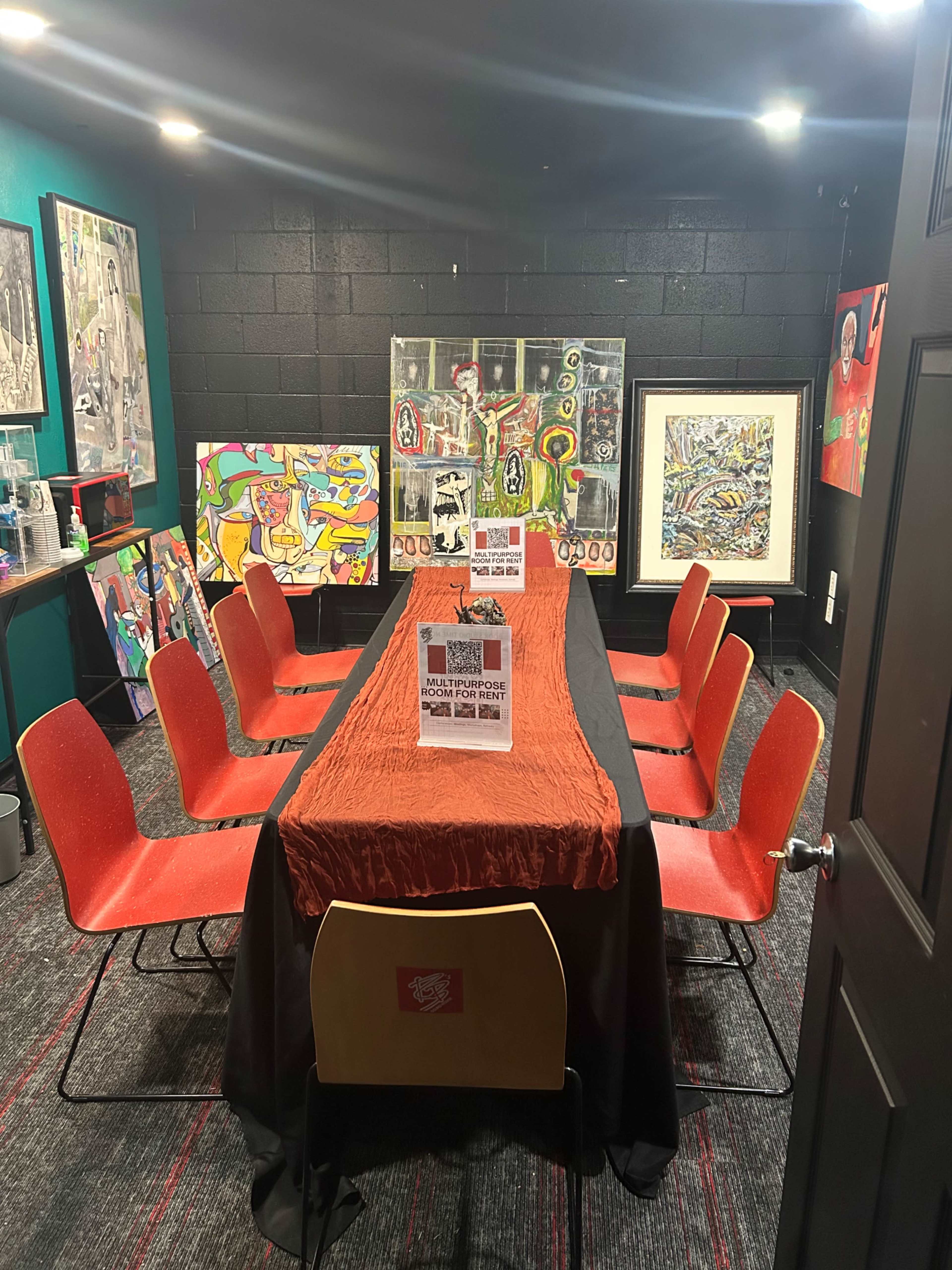 The image shows a meeting room with a long table covered in an orange tablecloth, surrounded by red chairs and adorned with colorful artwork on the walls.