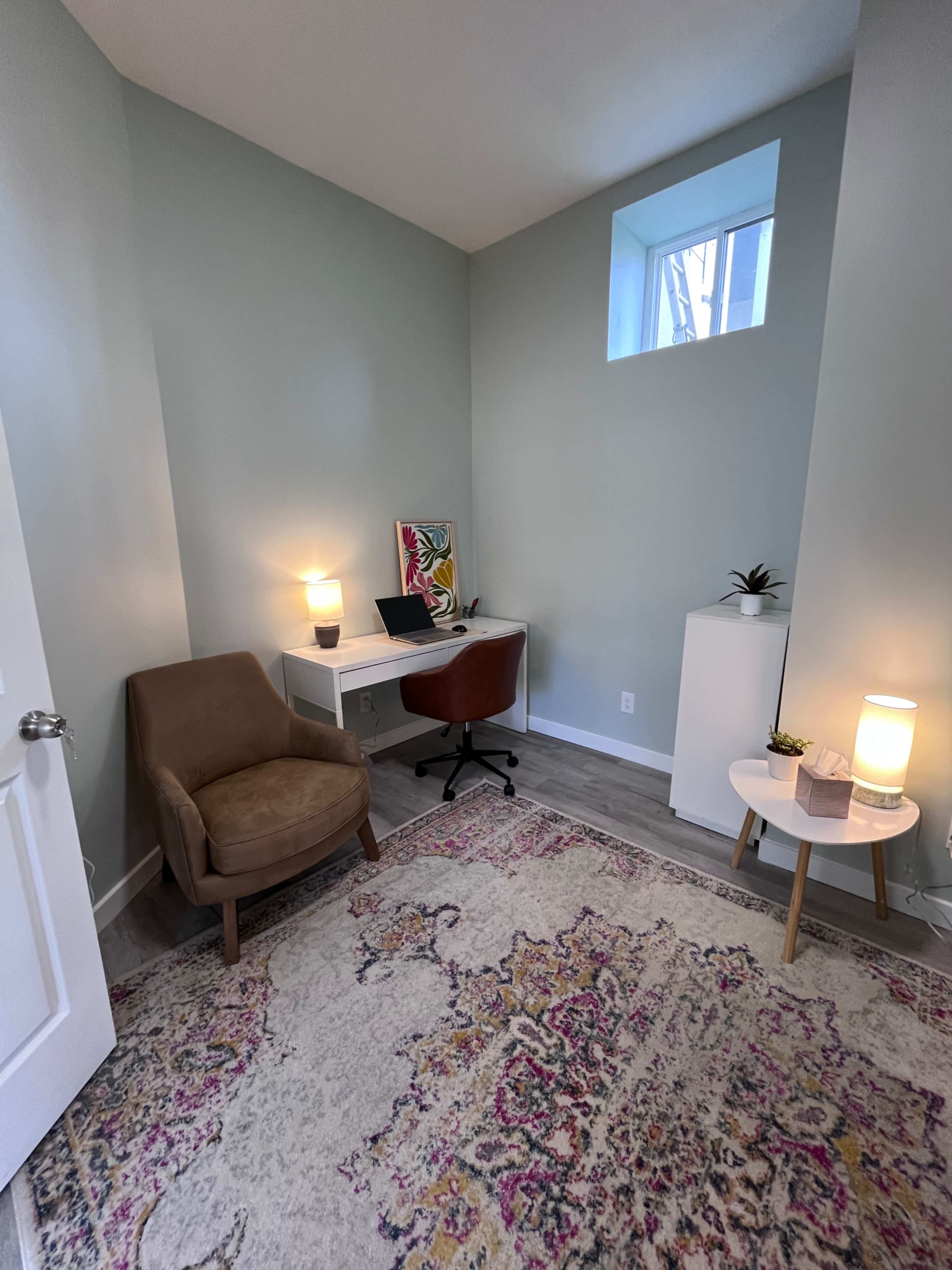 The image shows a small, well-lit office space featuring a desk with a computer, a chair, a decorative rug, and a small side table with a lamp.