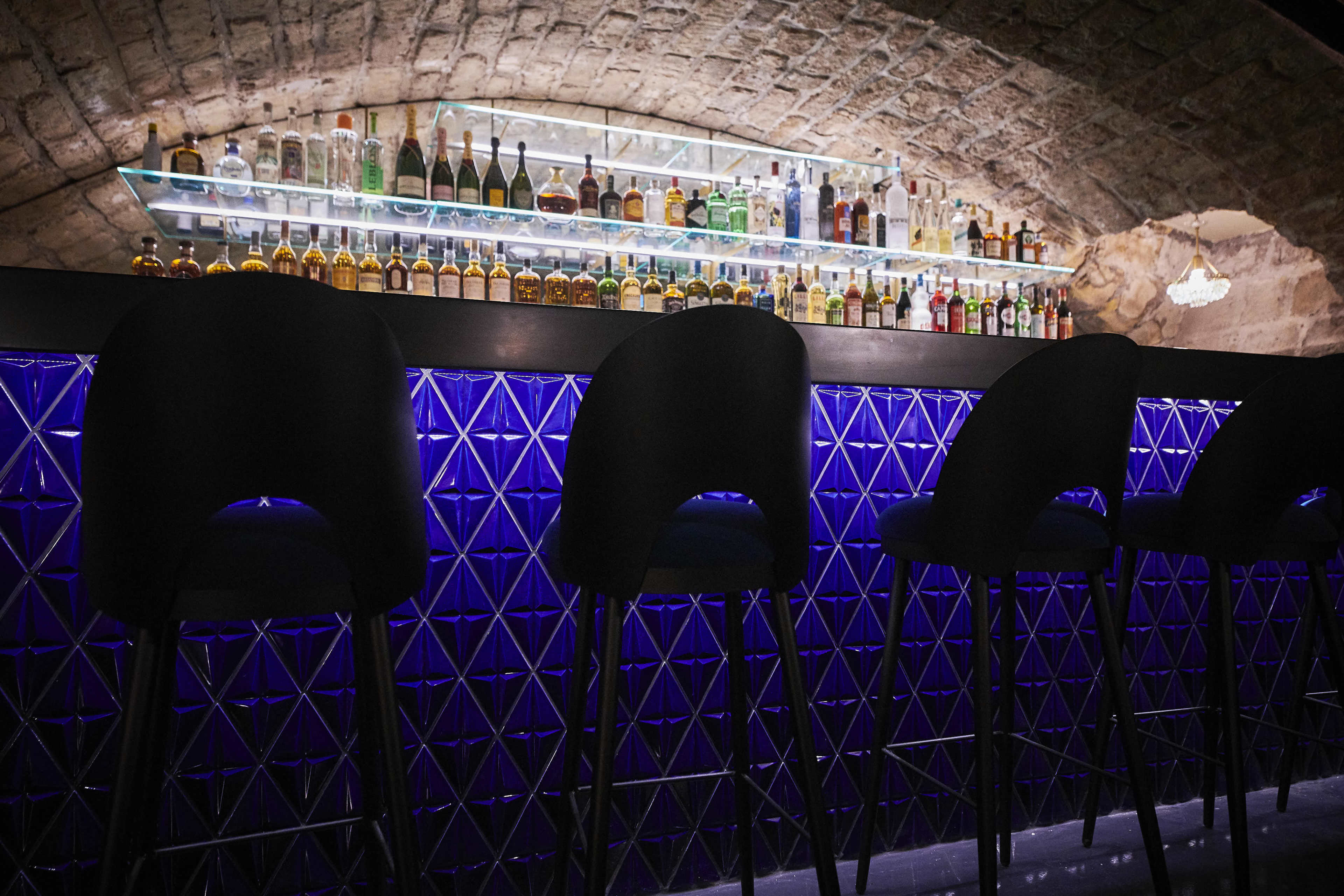 A dimly lit bar features a backlit shelf stocked with various bottles of liquor, accompanied by high black stools arranged in front of a blue geometric-patterned bar surface.