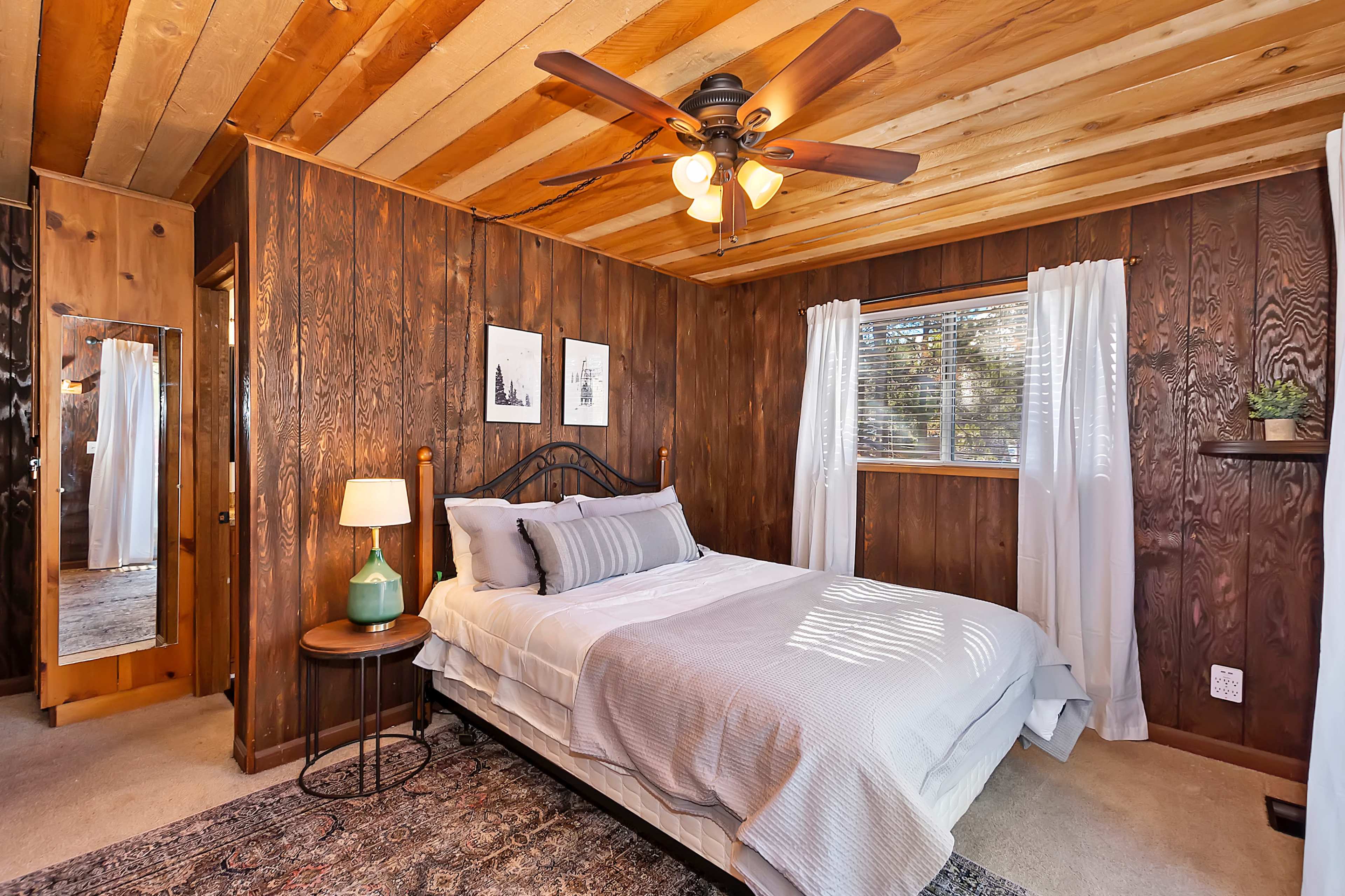 A cozy bedroom features a bed with light bedding, a wooden ceiling, and dark wood-paneled walls, along with a mirror and a window with white curtains.