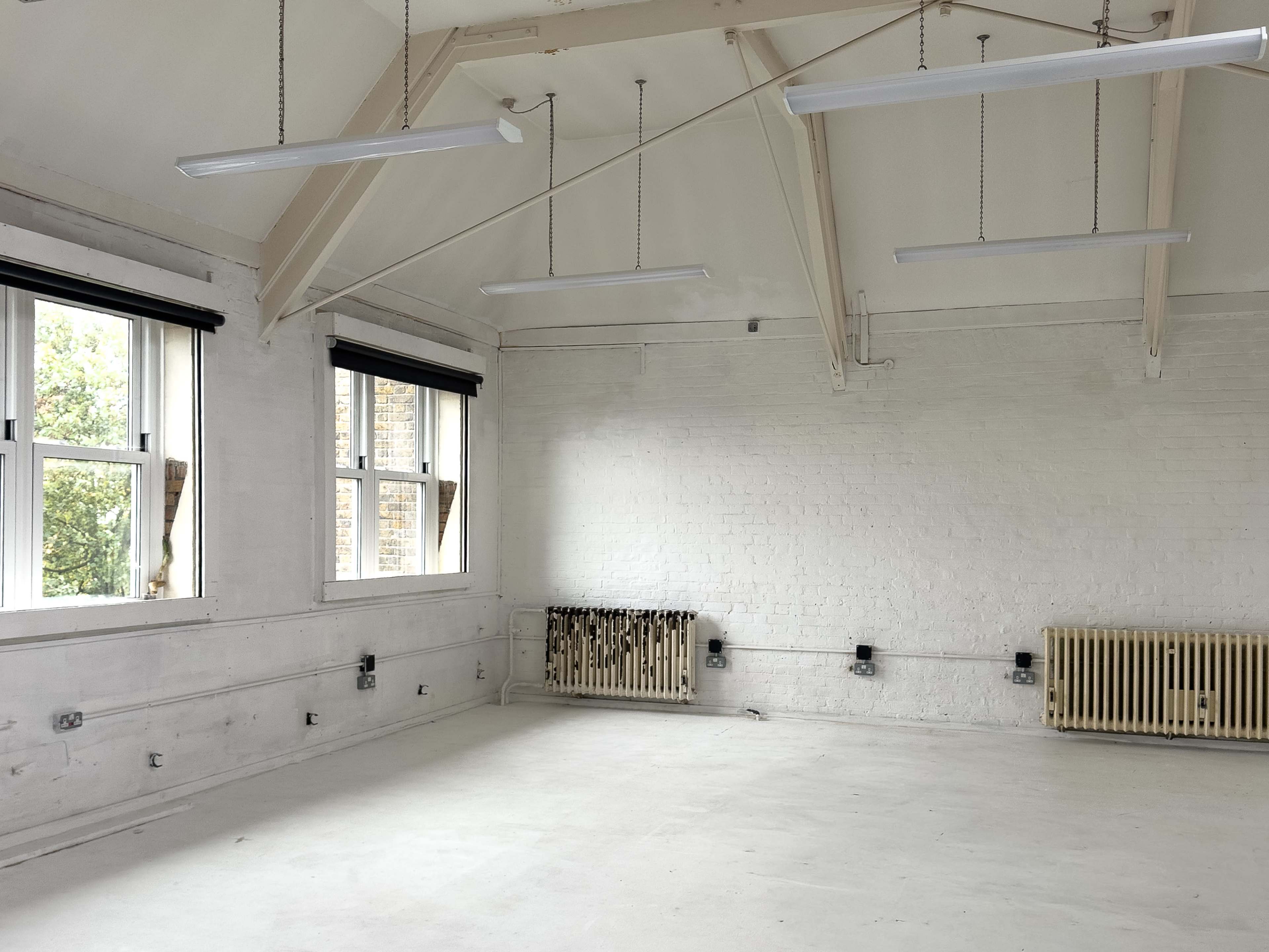 The image shows a vacant room with white walls, large windows, and overhead lighting fixtures.