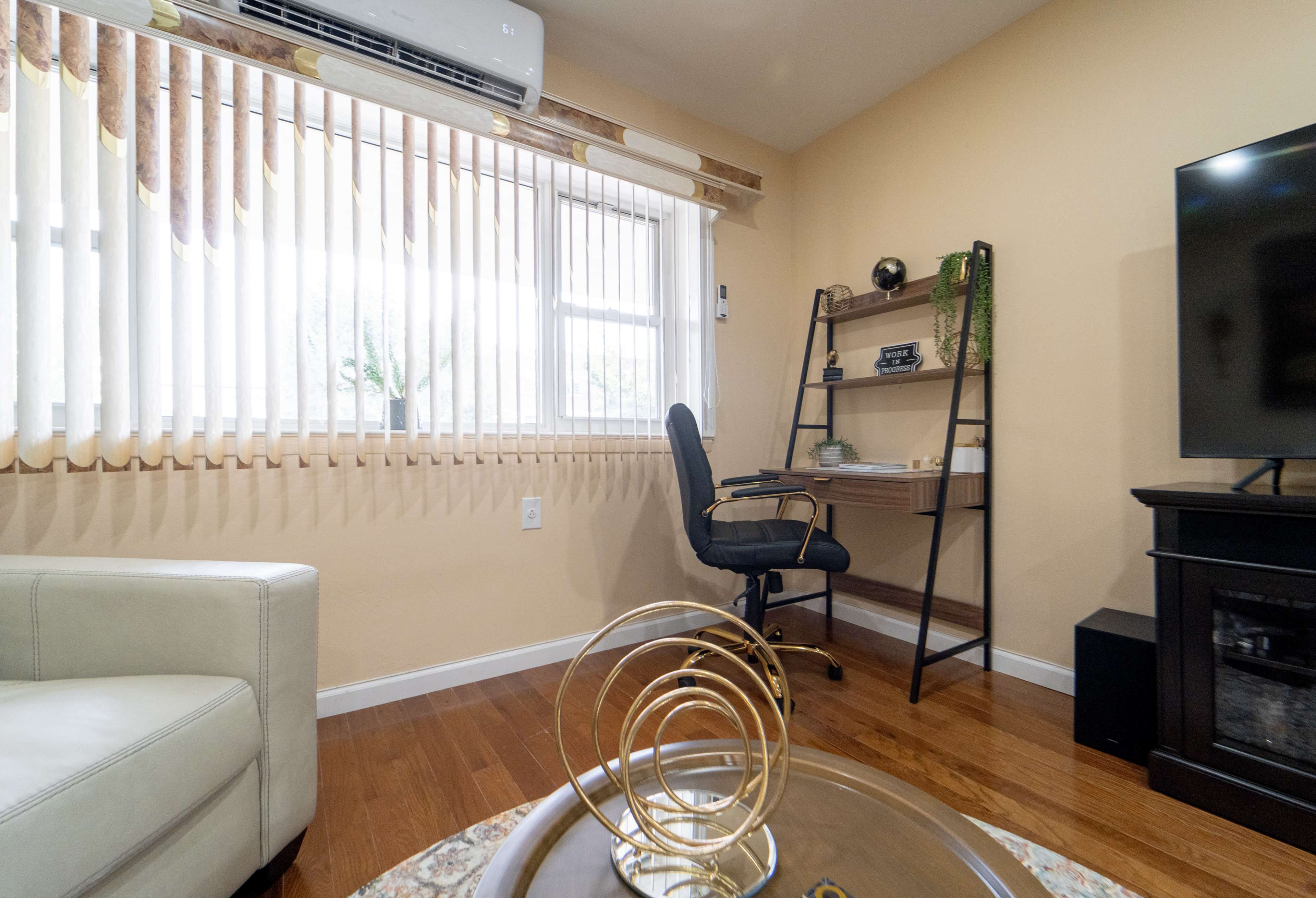 A cozy room features a window with vertical blinds, a desk with a chair, and a television stand.
