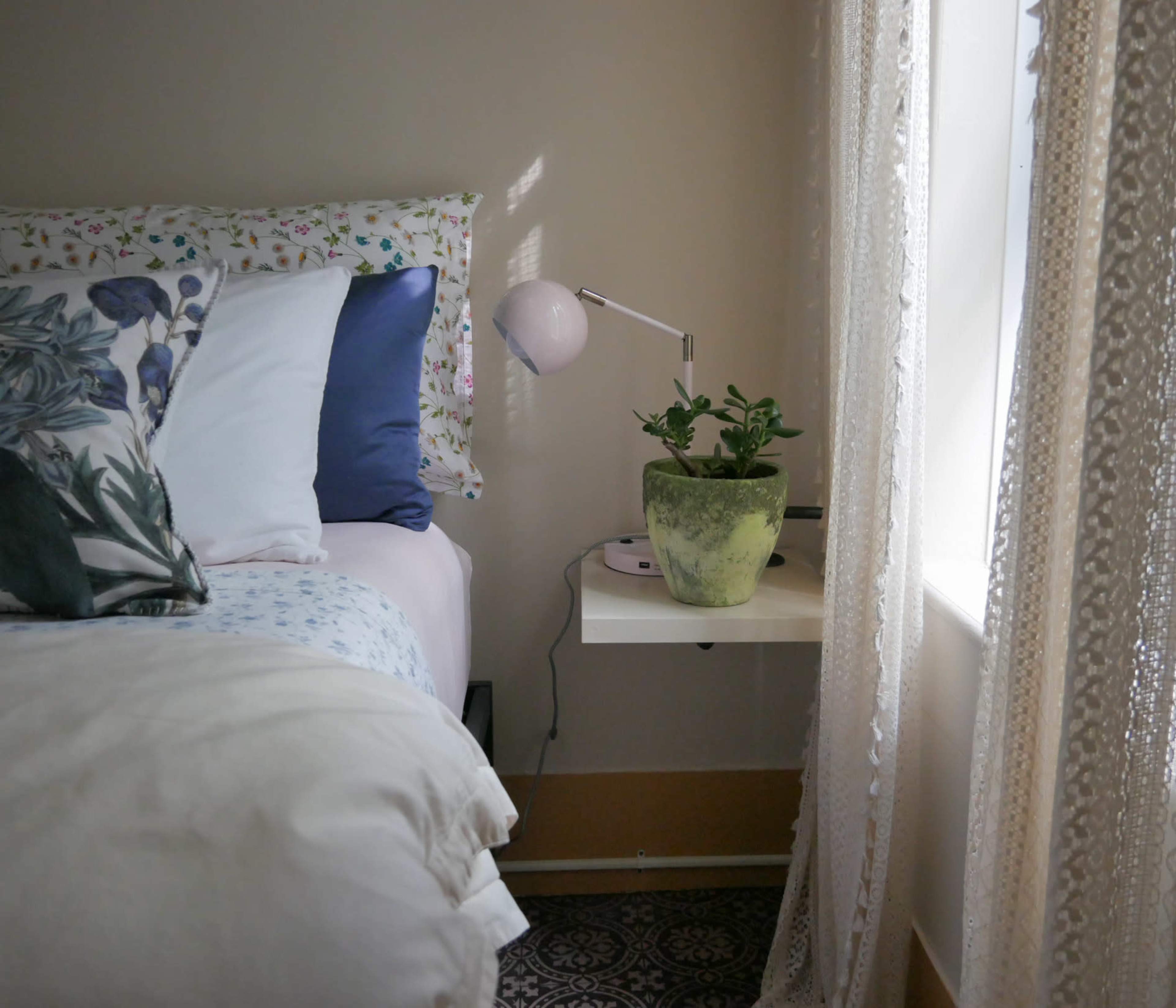 A neatly made bed with colorful pillows is positioned next to a small table containing a potted plant and a lamp, illuminated by natural light from a nearby window.