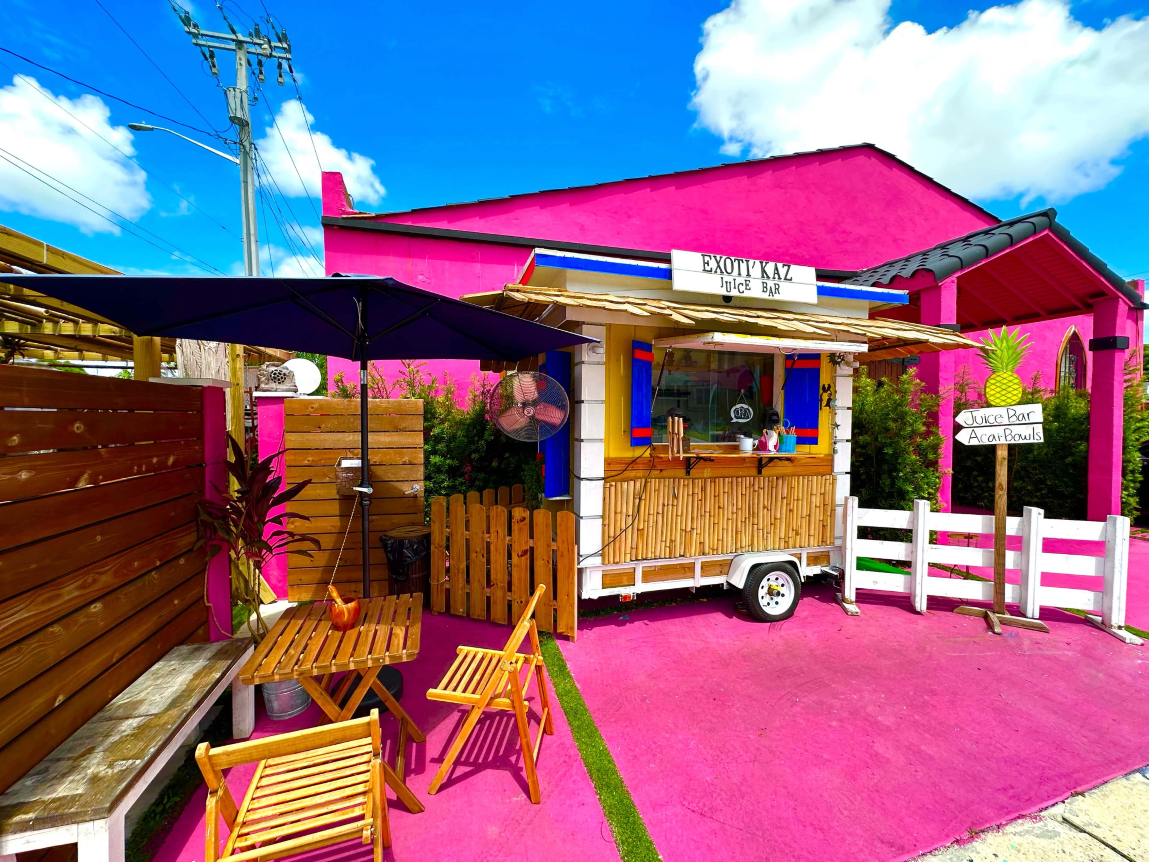A colorful food truck with a wooden serving counter and outdoor seating is set against a bright pink building.