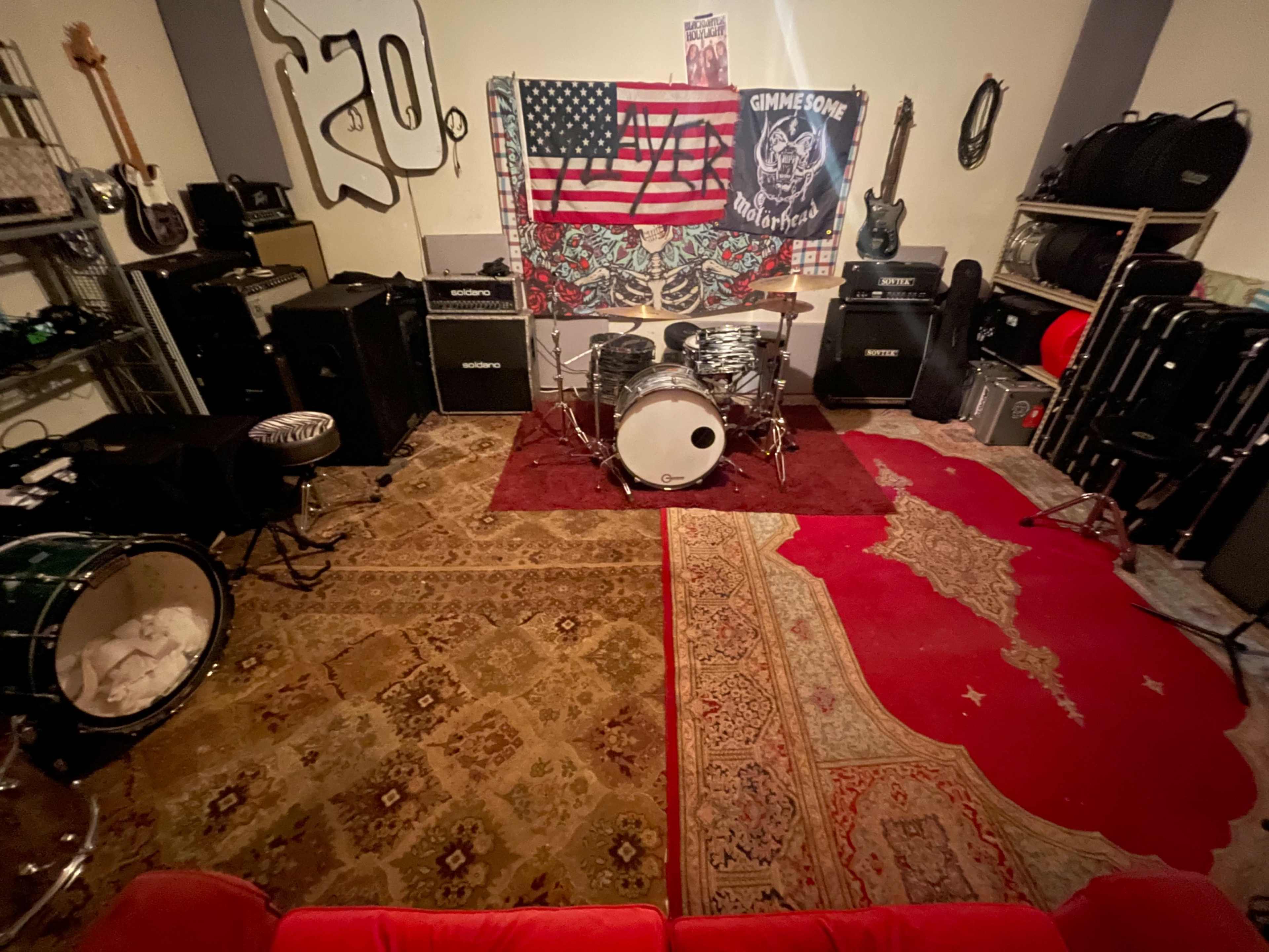 The image shows a music rehearsal space featuring a drum set on a rug, surrounded by amplifiers, guitars, and decorative flags on the walls.