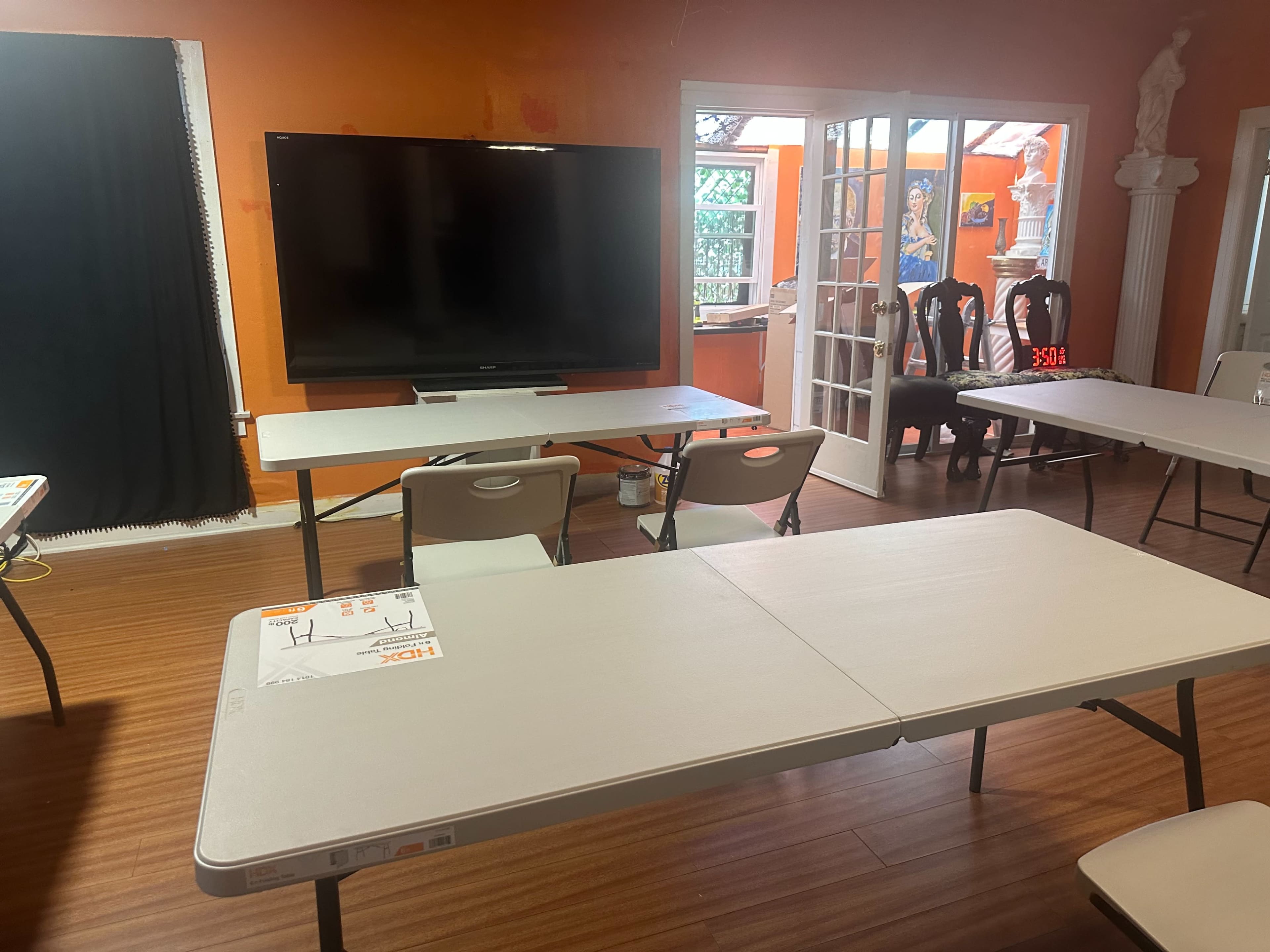 The image shows a room with multiple folding tables and chairs, a large television on the wall, and a view of an outdoor area through glass doors.