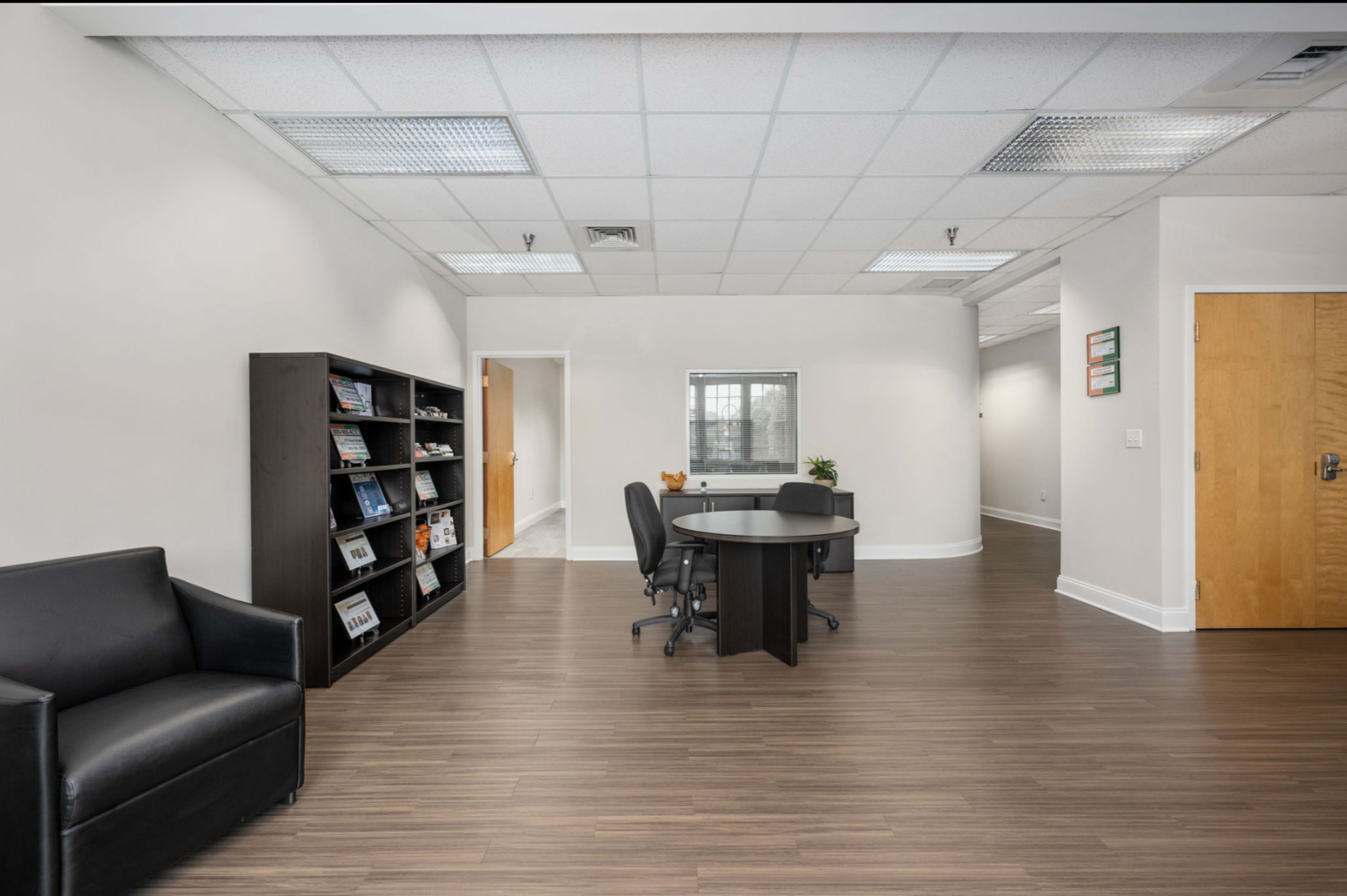 The image shows an office space with a dark wooden floor, a round table with chairs, a black sofa, and a bookshelf filled with various items.