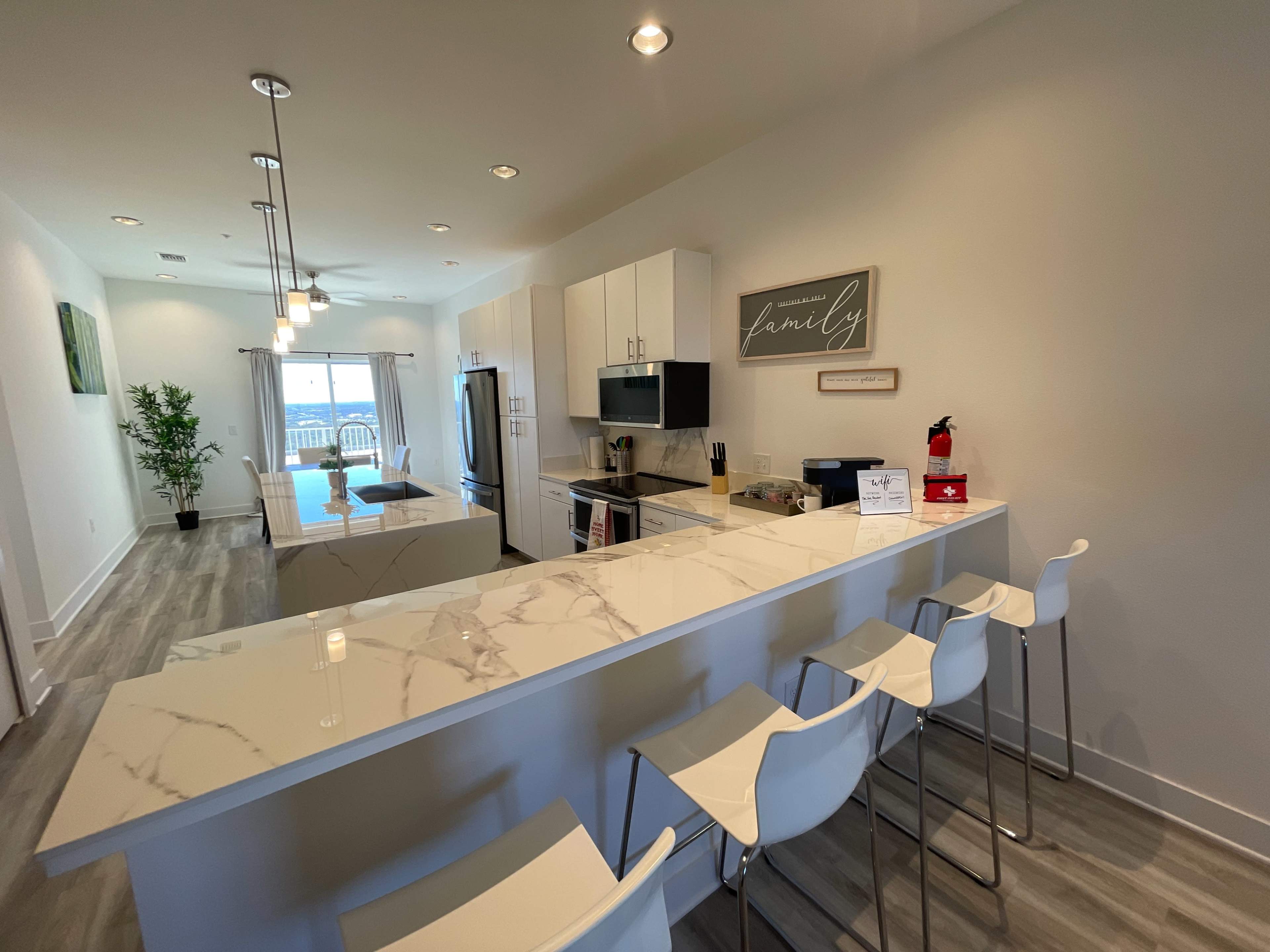 A modern kitchen with white cabinetry, a marble countertop, and bar stools, leading into a living area with a view of a balcony.