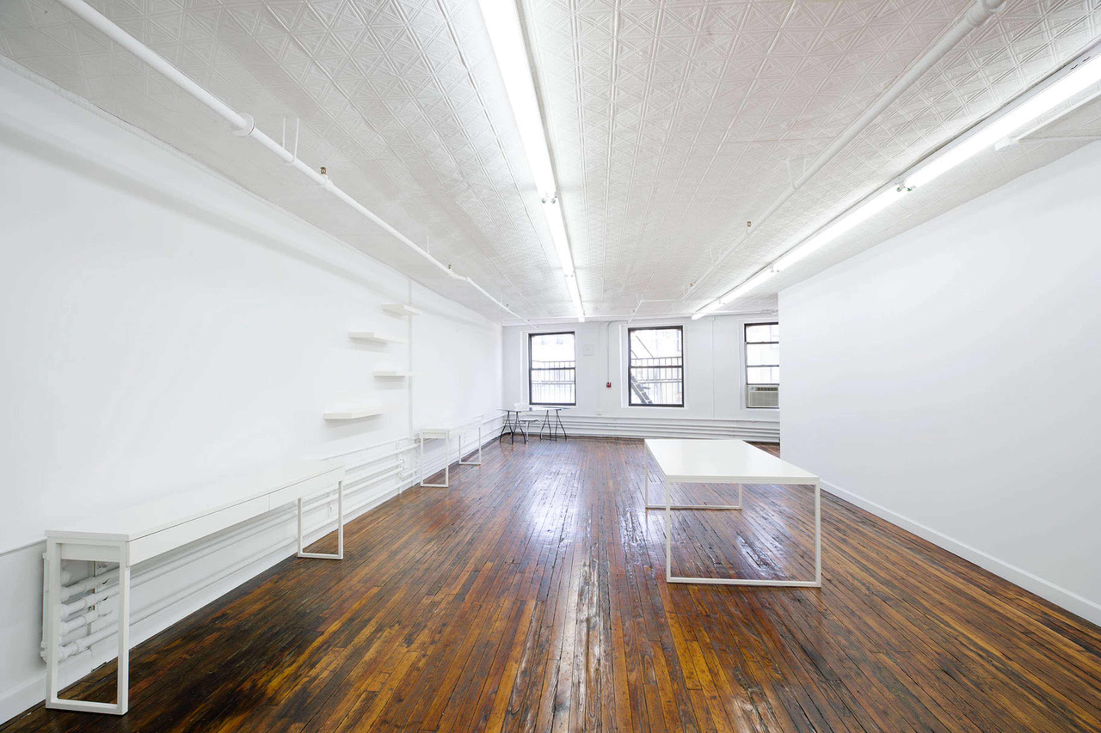 The image shows a spacious, empty room with wooden floors, white walls, and minimal furnishings, including a table and shelves.