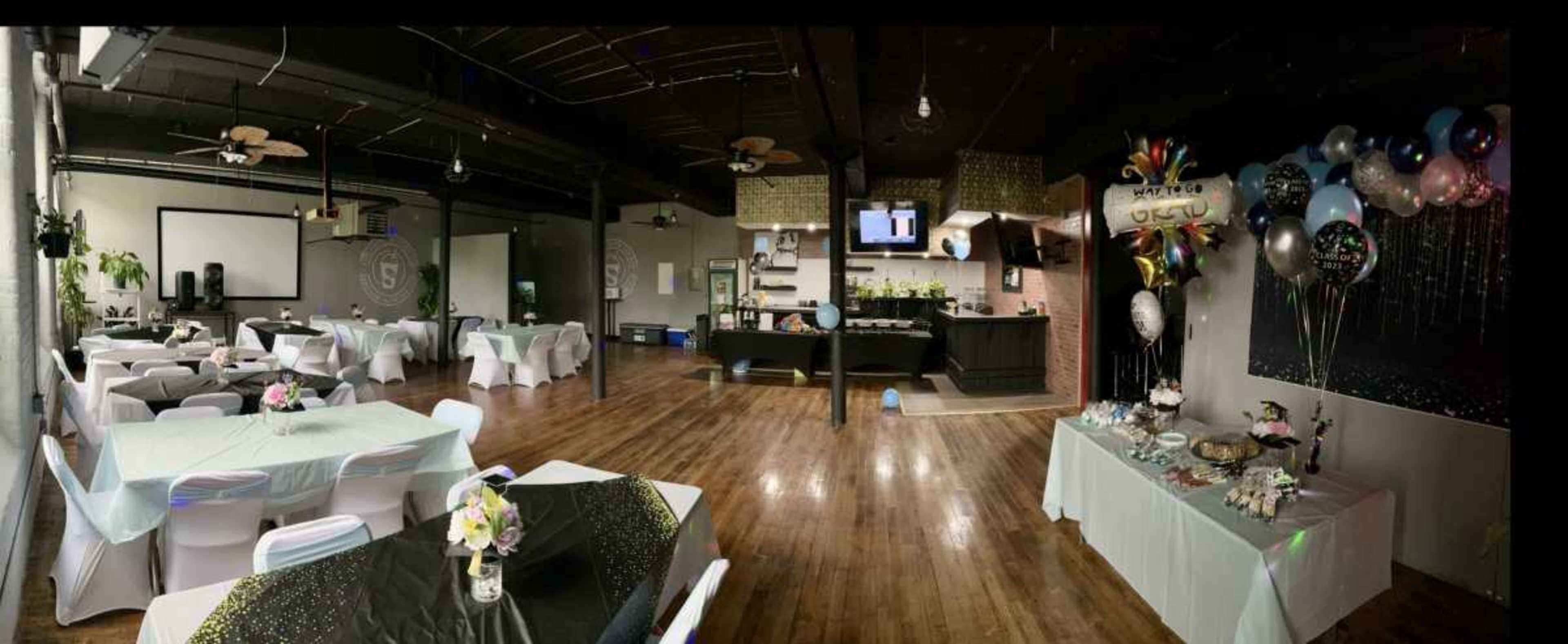 The image shows a spacious event venue featuring tables adorned with decorative cloths and centerpieces, a buffet station, and a festive balloon display along one wall.