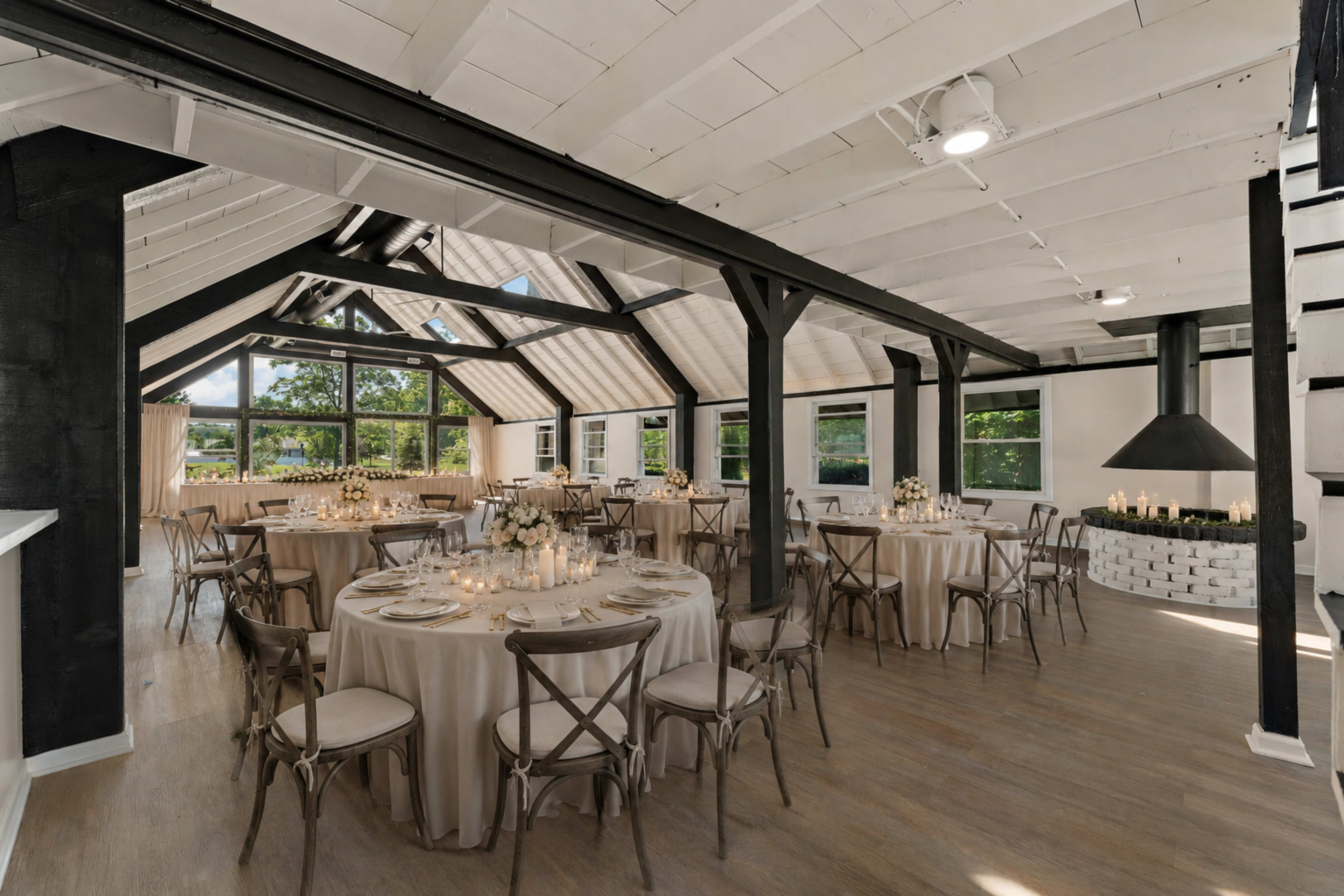 The image shows a well-lit event space featuring round tables with cream-colored tablecloths and wooden chairs, set under a vaulted ceiling with large windows and a modern fireplace.