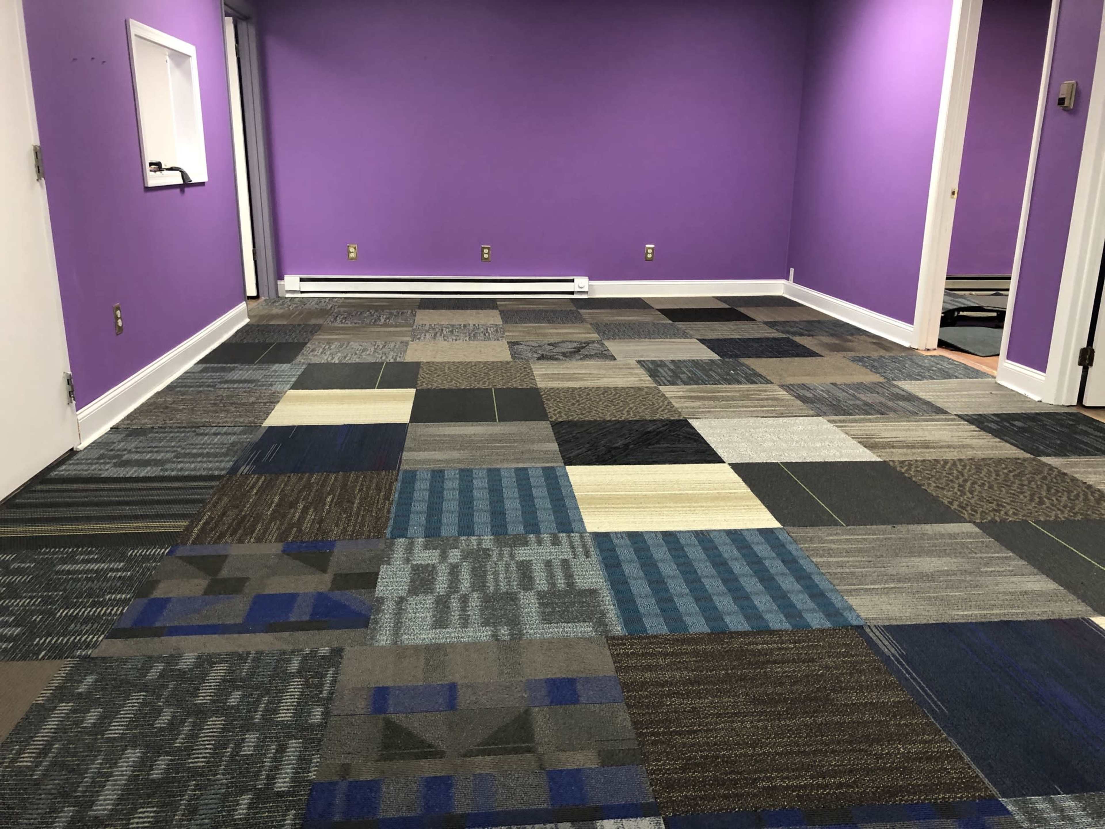 An empty room with a purple accent wall and a floor featuring a patchwork of various carpet tiles in different colors and patterns.