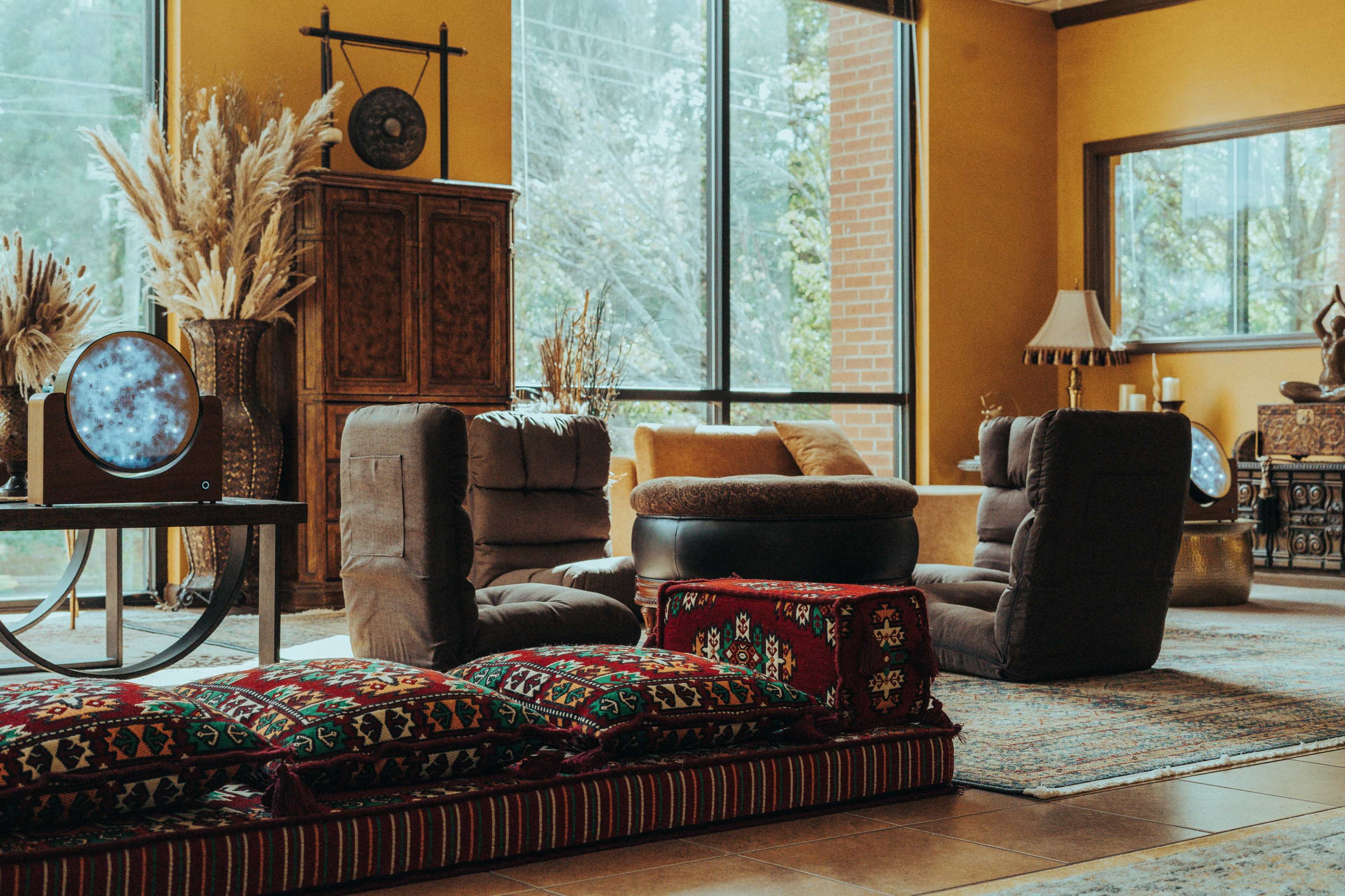 The scene features a spacious living room with large windows, two comfortable armchairs, a round ottoman, and decorative pillows arranged on the floor.