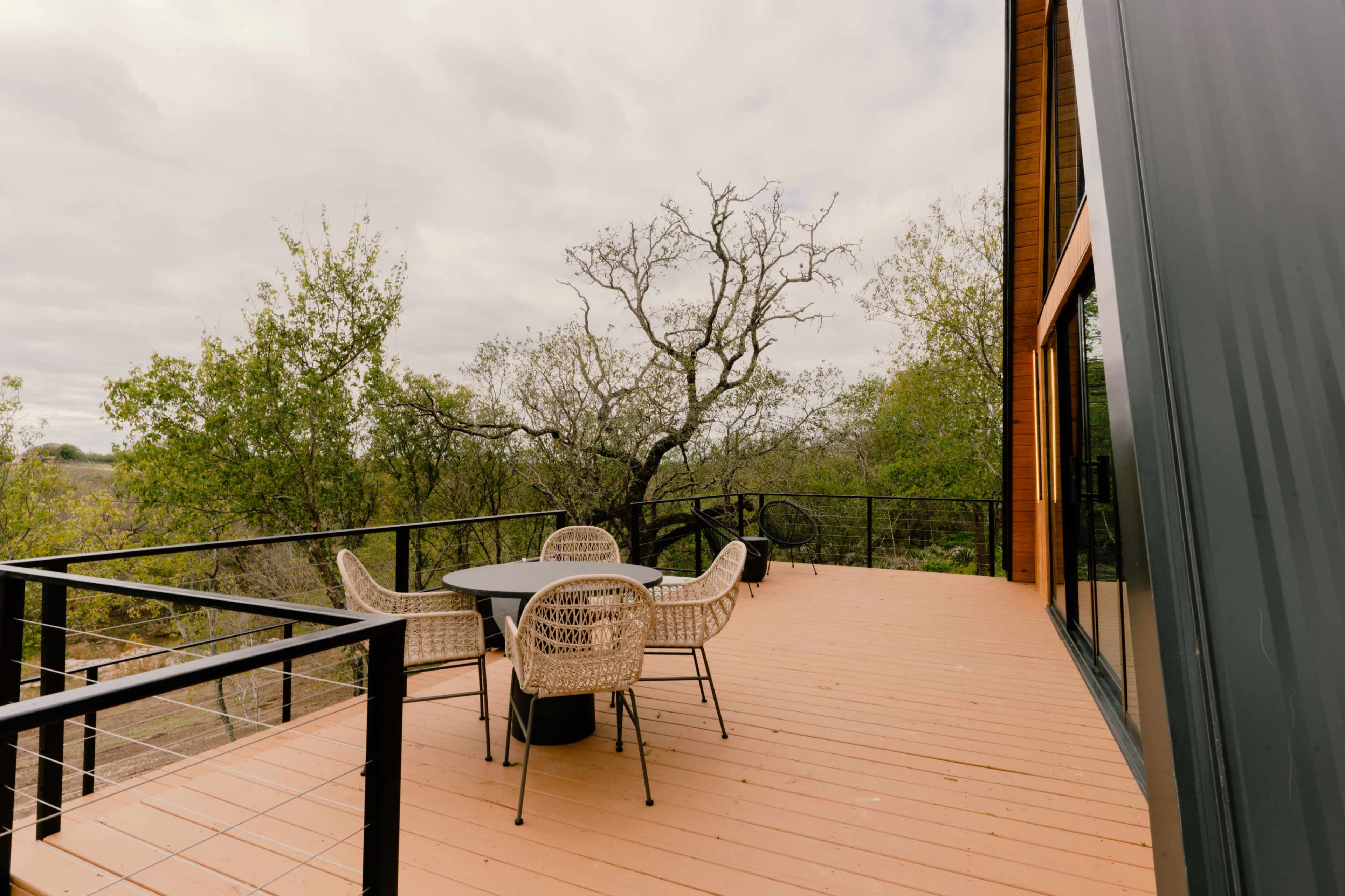 A wooden deck features a round table and four woven chairs, surrounded by trees under an overcast sky.